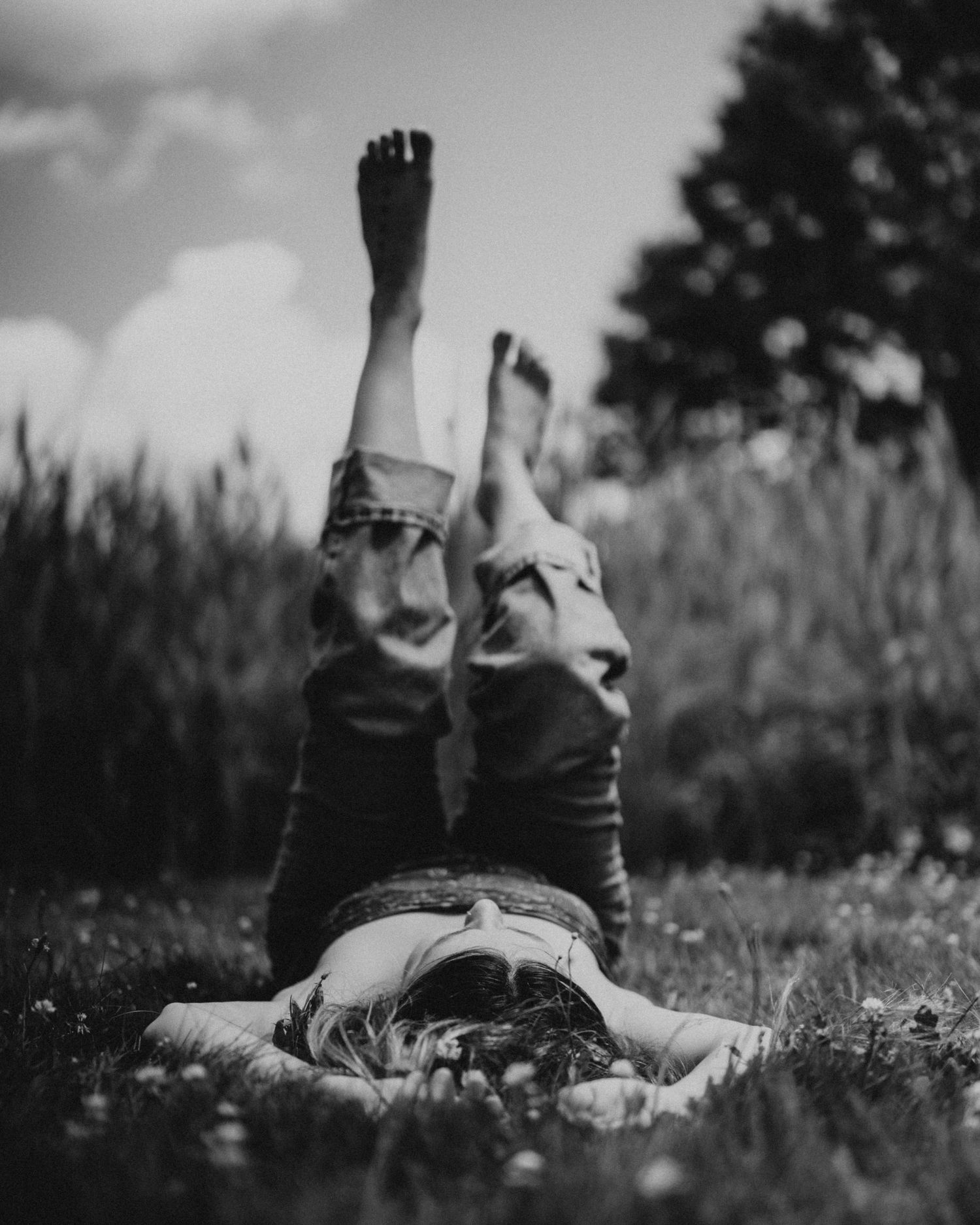 Black and white photo of someone lying in grass with legs raised against a cloudy sky, creating a dreamy summer mood.