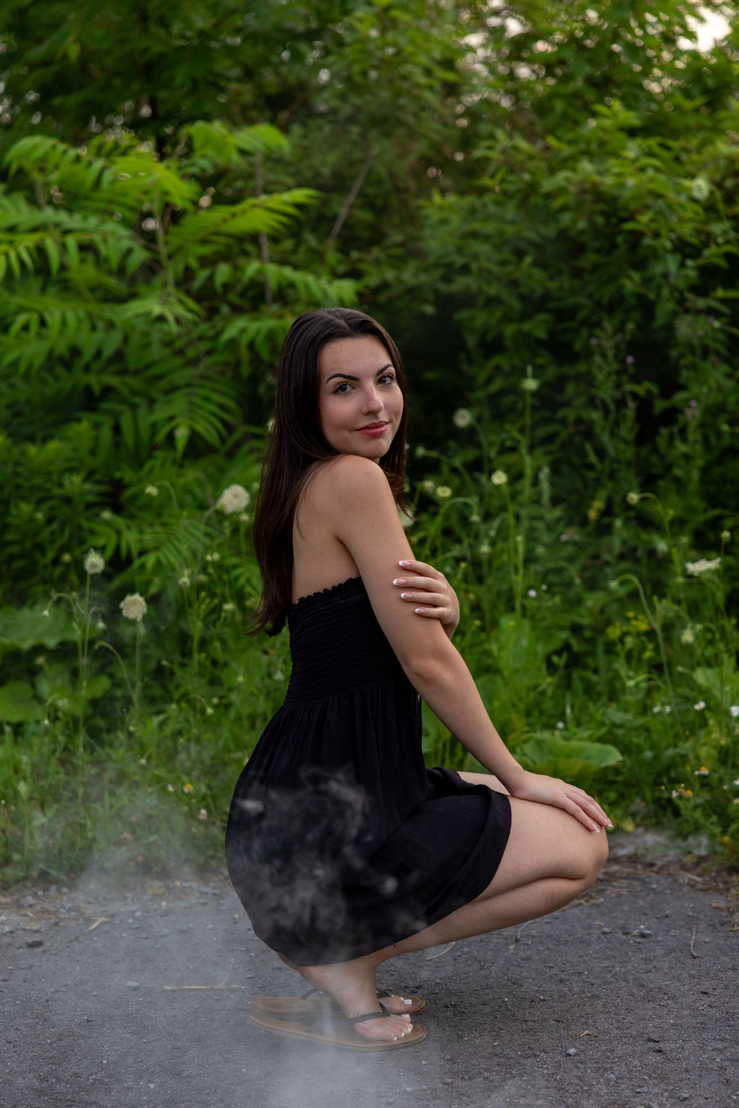 A woman in a black dress poses gracefully on the ground against a lush green forest backdrop.