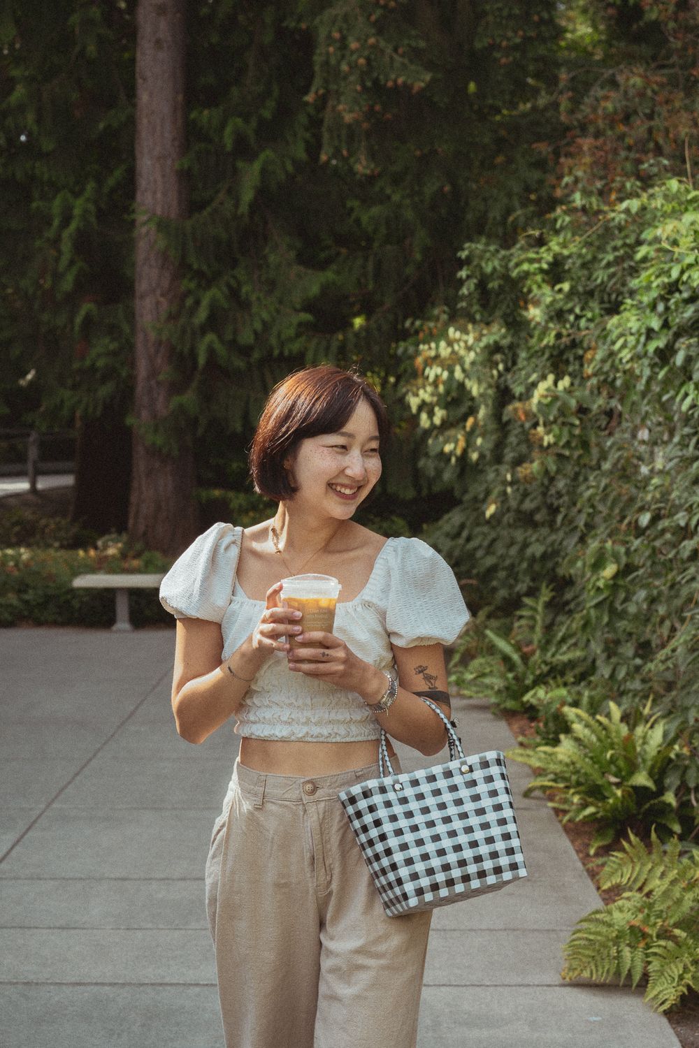 Woman in white crop top and beige pants enjoys a meal while carrying a checkered handbag in an outdoor setting.