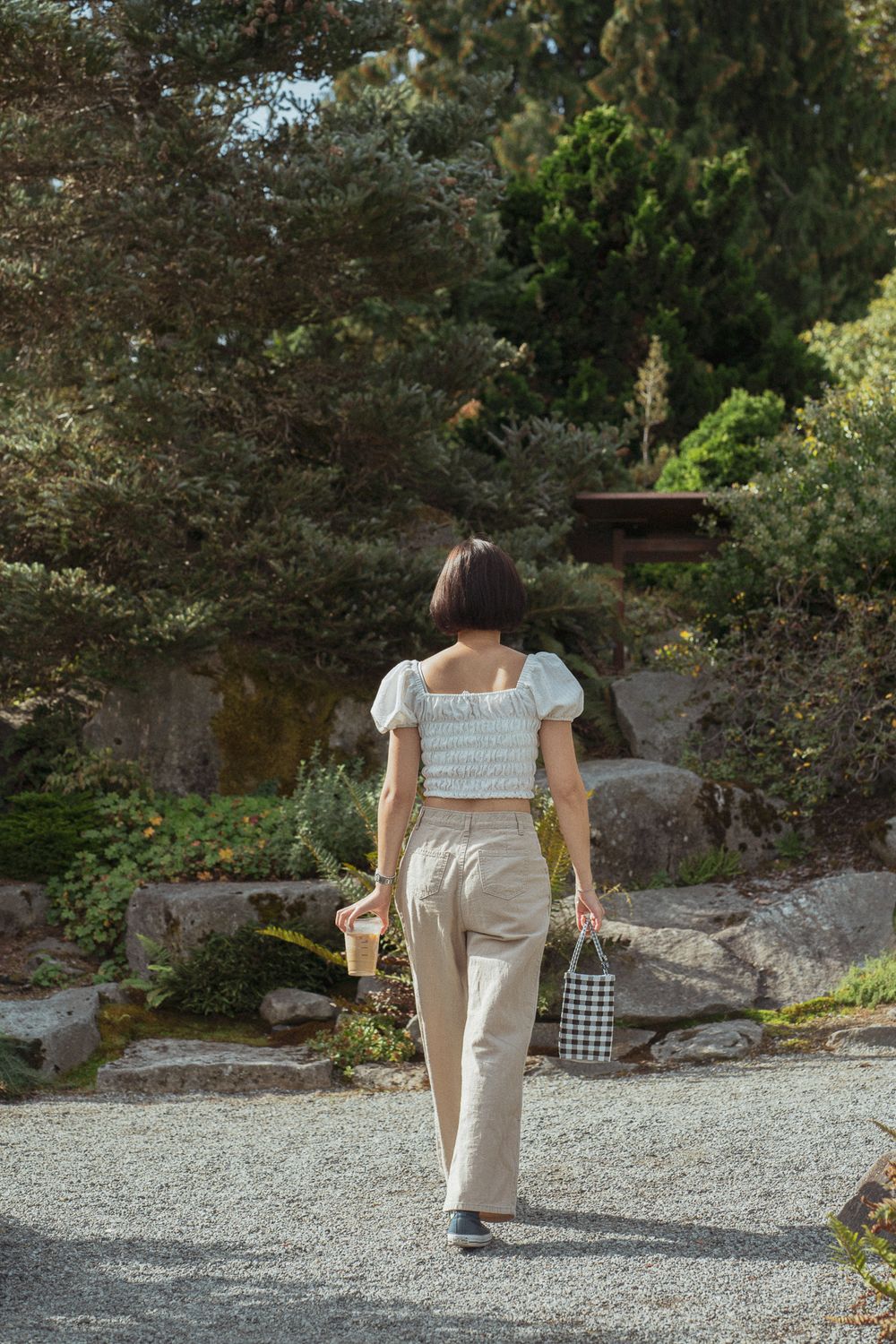 Woman in light colored pants and top walking on gravel path through a garden setting.