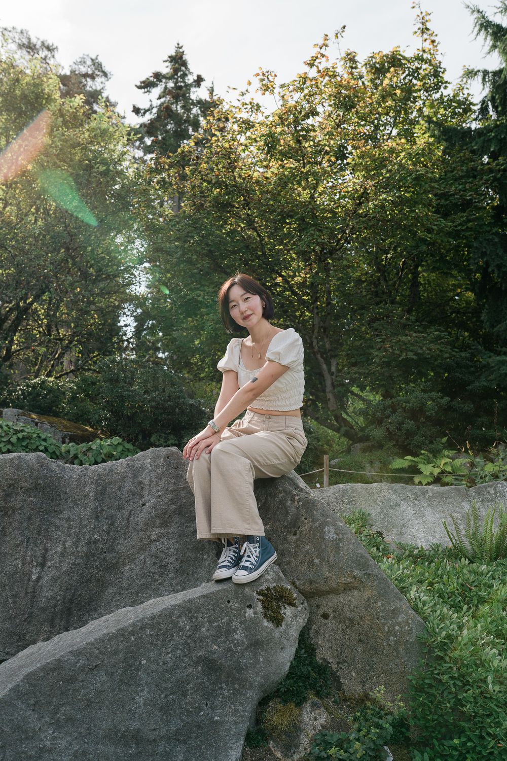 Person in white dress sitting on large gray boulder in natural outdoor setting surrounded by greenery and trees.