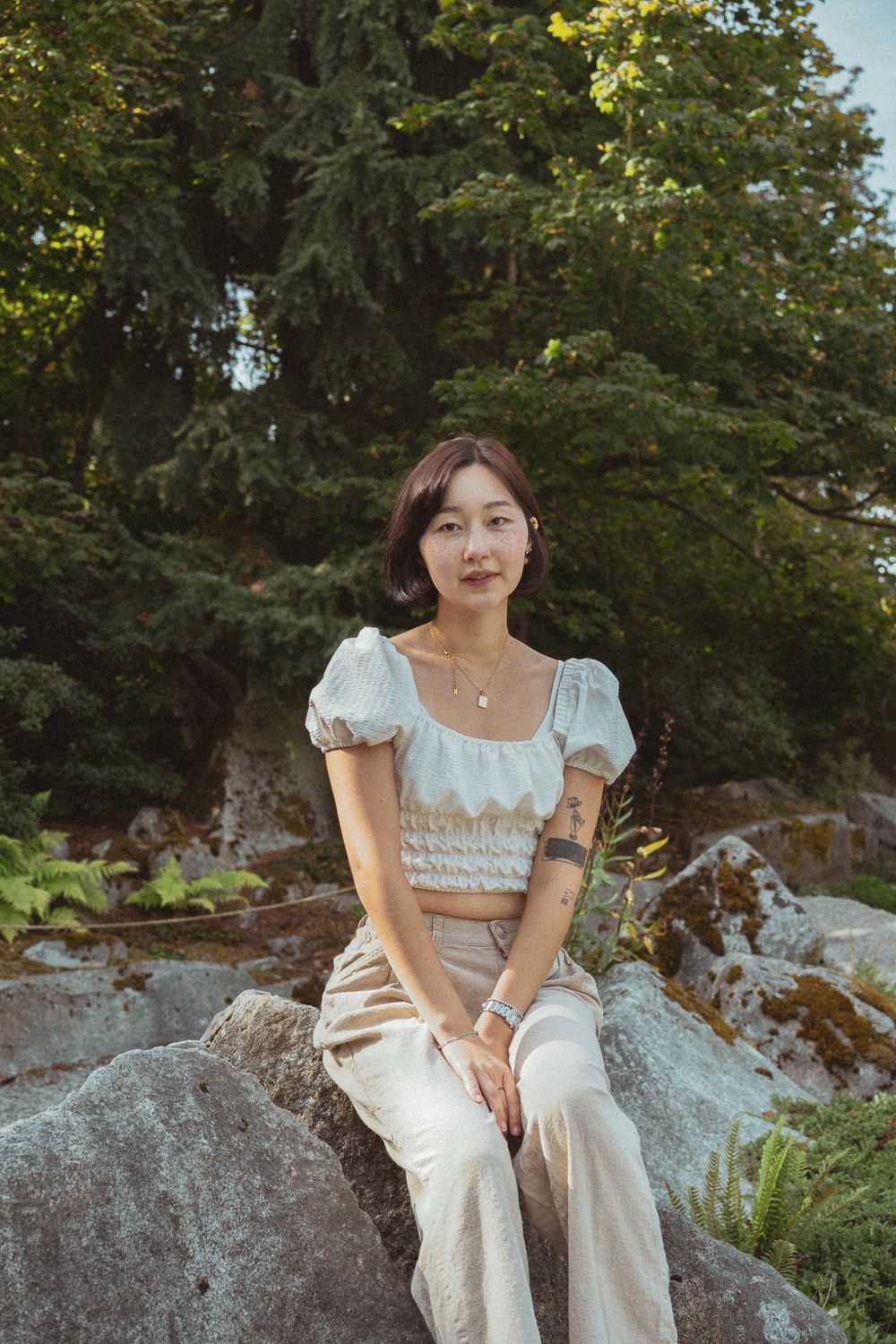 A person in a white puff sleeve top sits on a large rock in a natural outdoor setting with lush greenery.