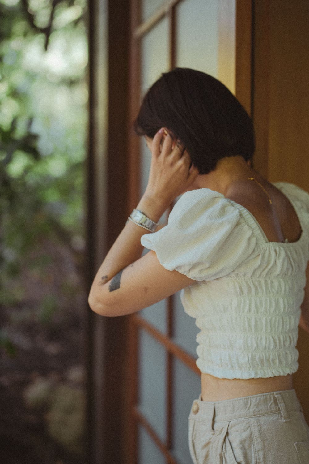 Person in a white smocked crop top and light pants stands near a window, photographed from behind in soft lighting.