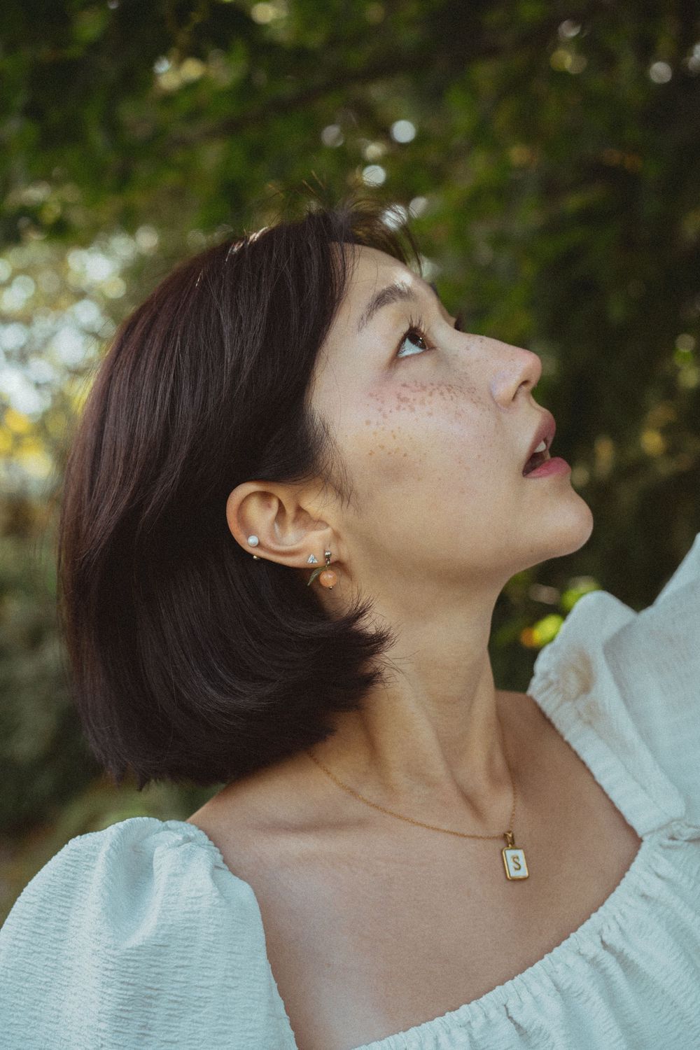 A profile view against a green background showing a gentle upward gaze while wearing a white blouse and delicate necklace.