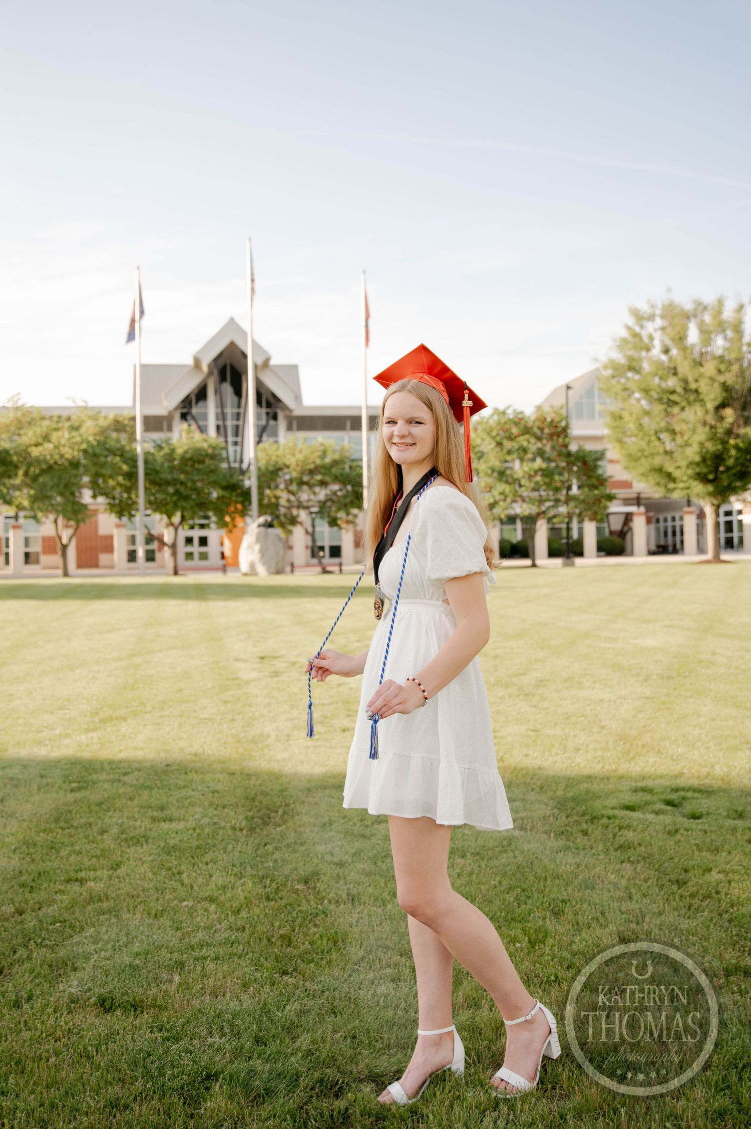 Graduate in white dress and red cap poses for outdoor portraits with diploma on sunny grassy campus lawn.