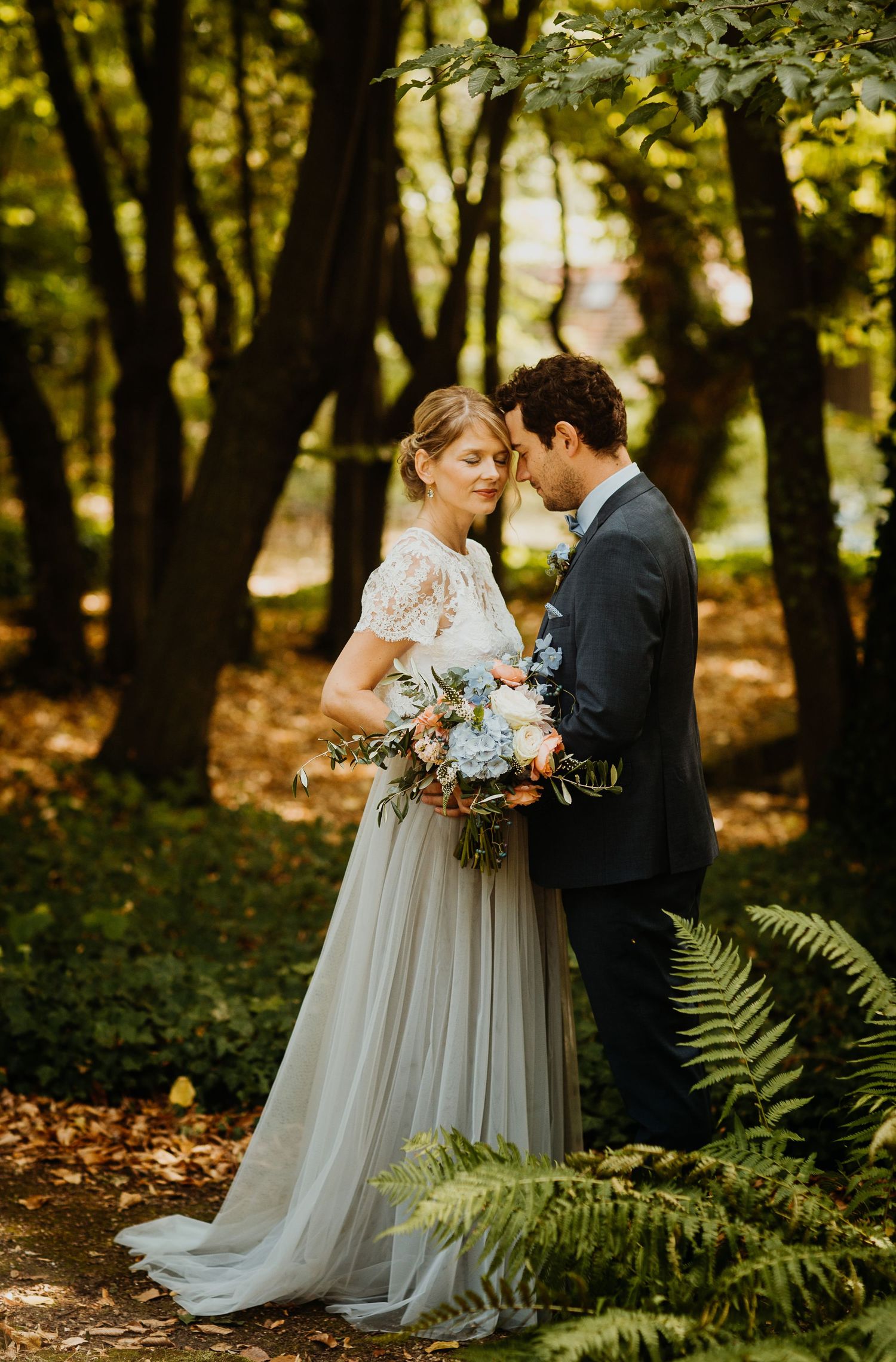 A couple in wedding attire shares an intimate moment in a lush wooded forest setting with sunlit trees and ferns.