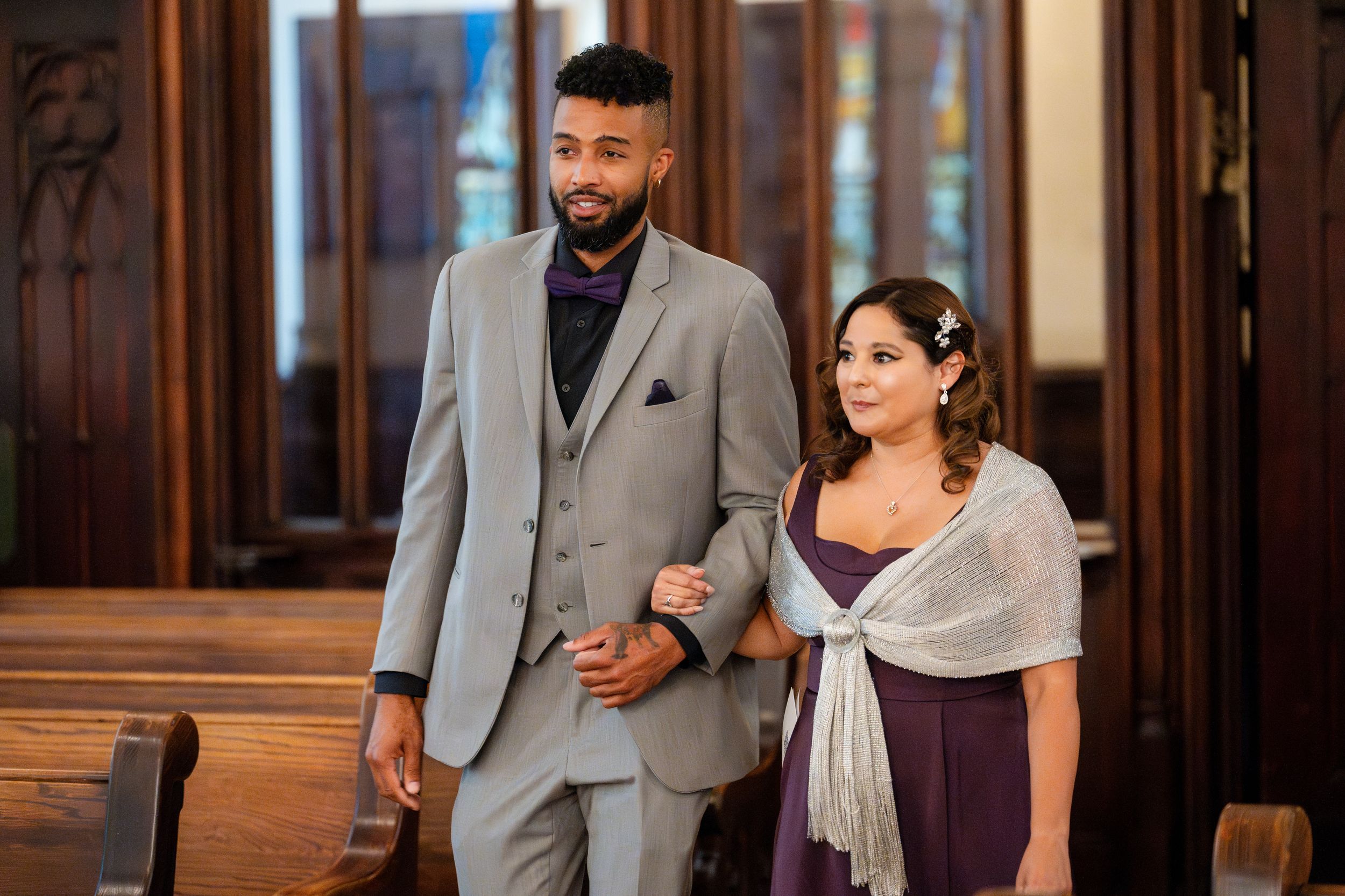 Well-dressed couple in formal attire walking arm-in-arm through an elegant hallway with wooden paneling.