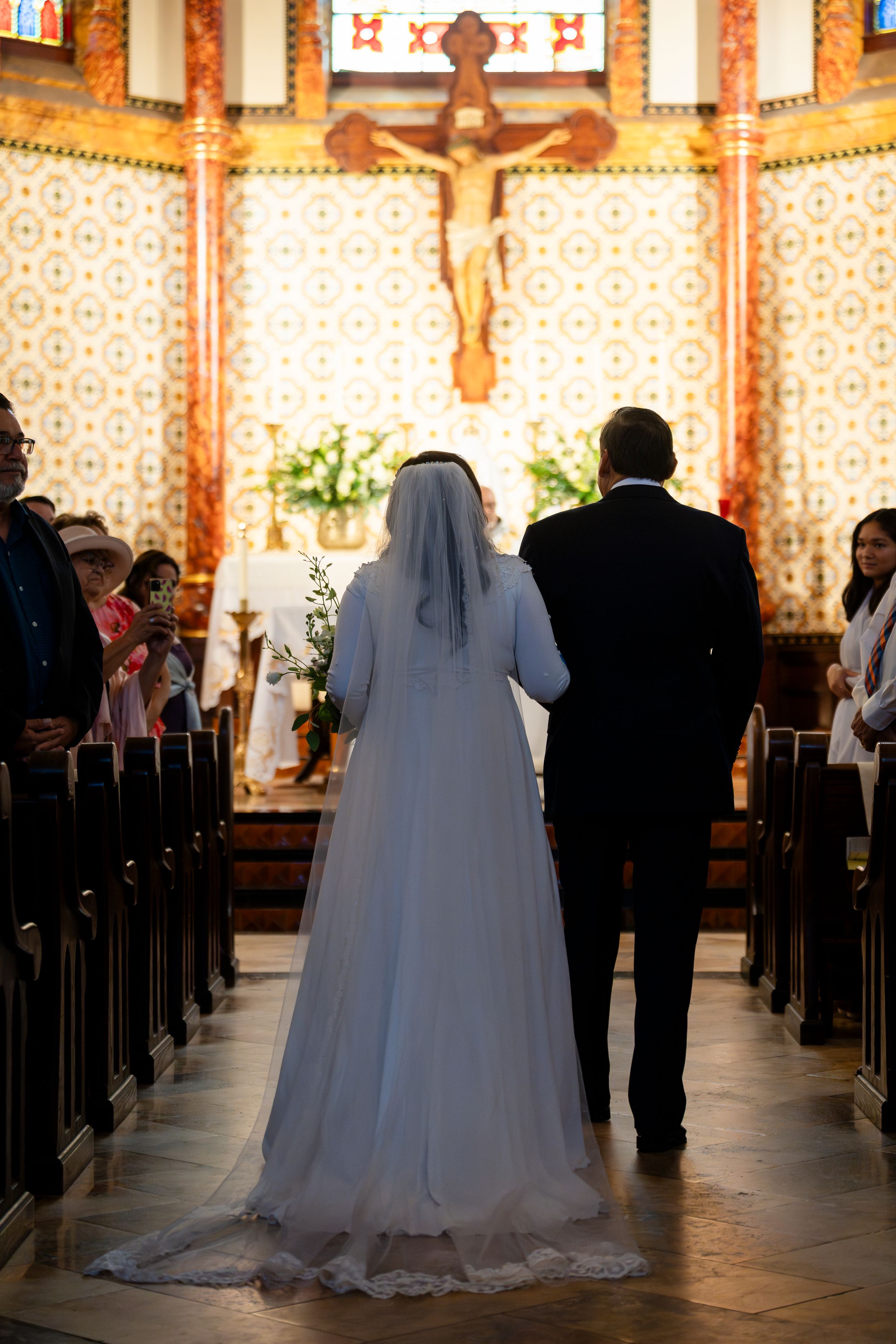 Bride and groom stand at altar during Catholic church wedding ceremony with crucifix and decorative wall patterns visible.