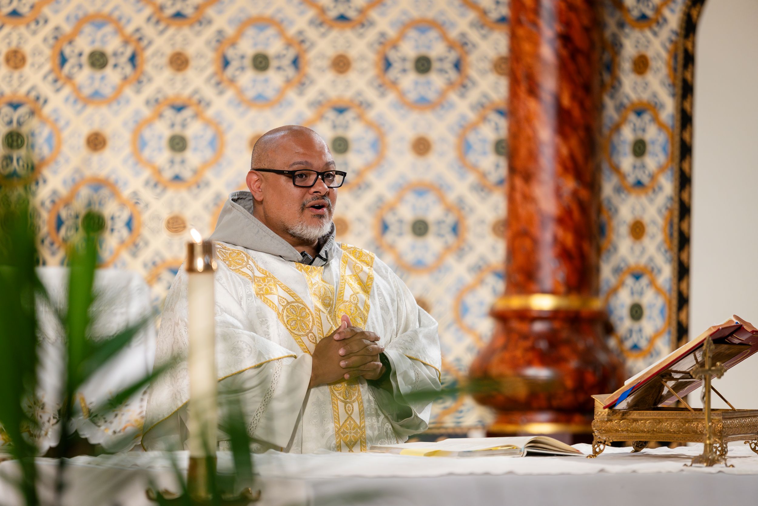 Clergy member wearing glasses and white and gold vestments smiles while leading service at ornate altar with tiled background.