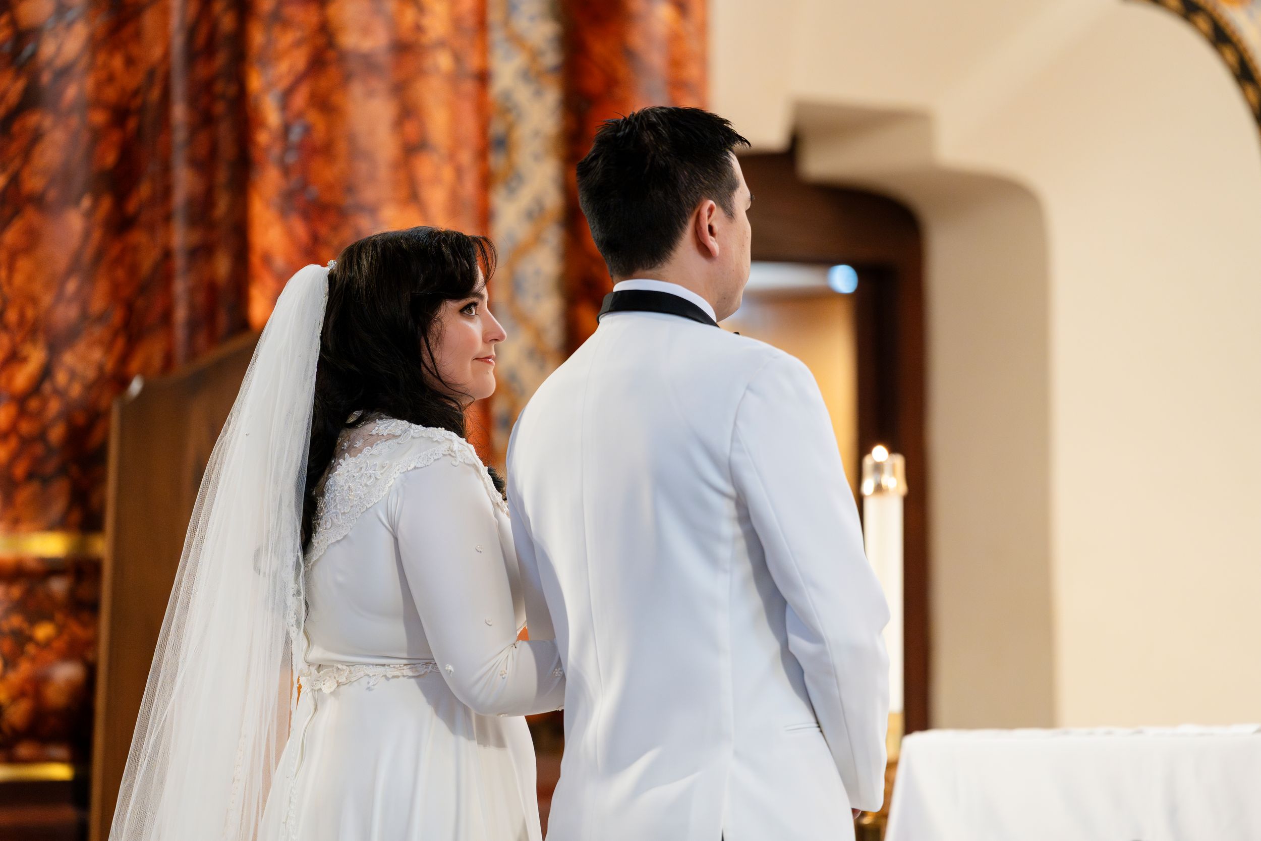 A couple in white wedding attire stand facing an altar with ornate orange and brown decor.