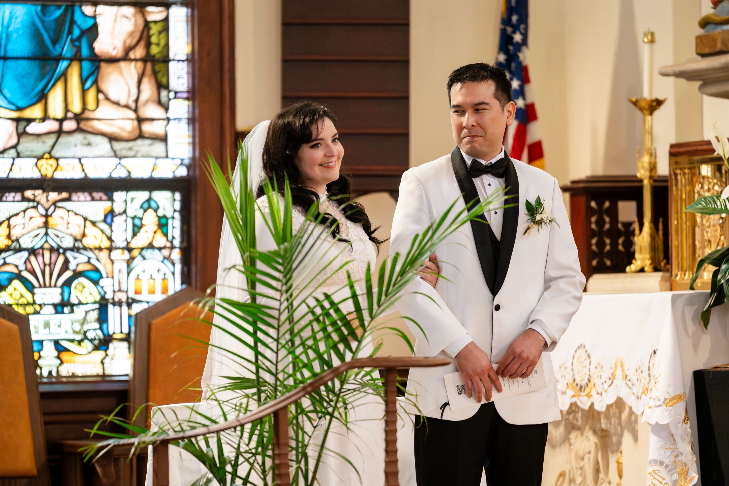 A couple stands together during their wedding ceremony in a church with stained glass windows and palm decor.