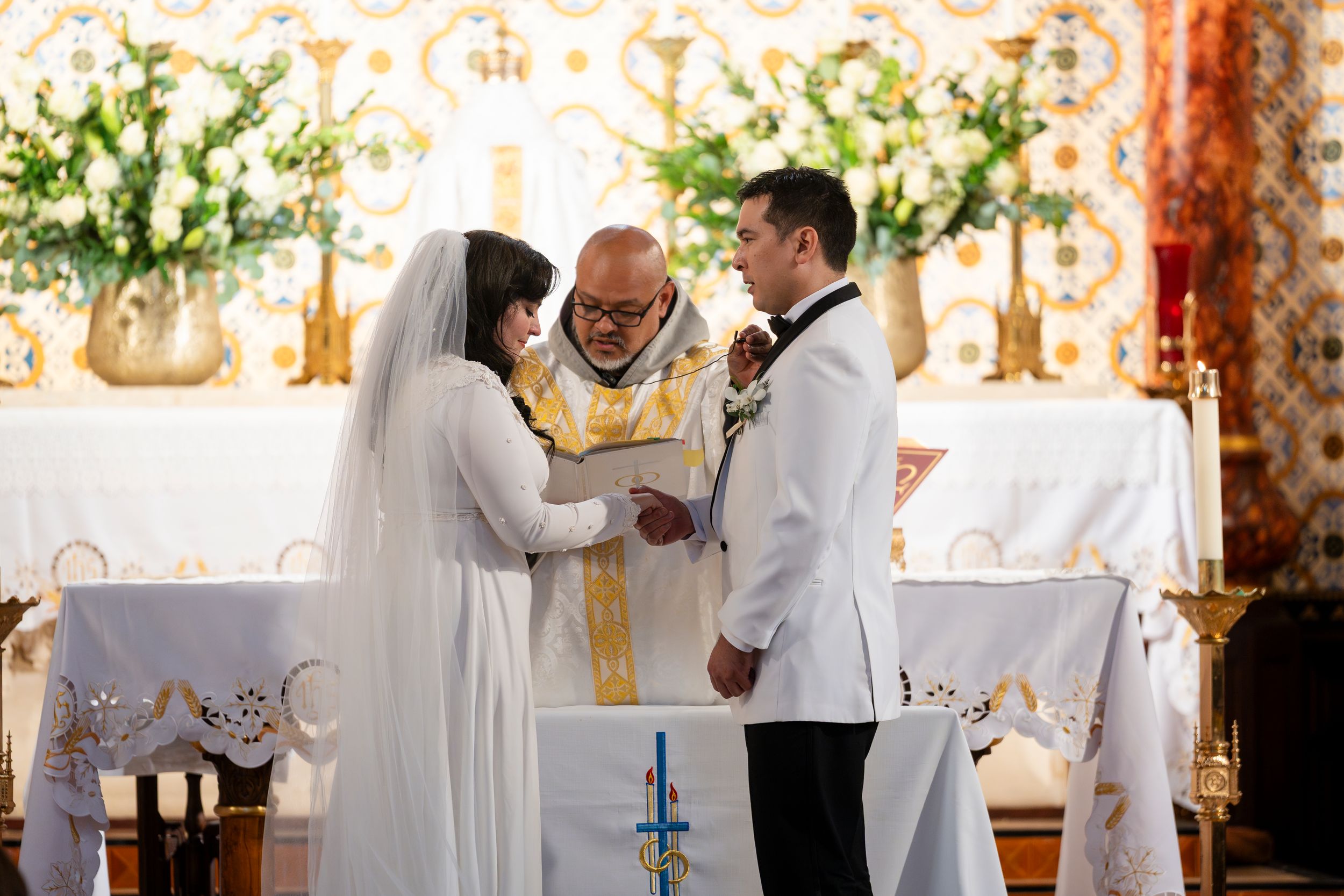 A wedding ceremony takes place inside a church with a clergy member officiating in gold robes as the couple exchanges vows at the altar.