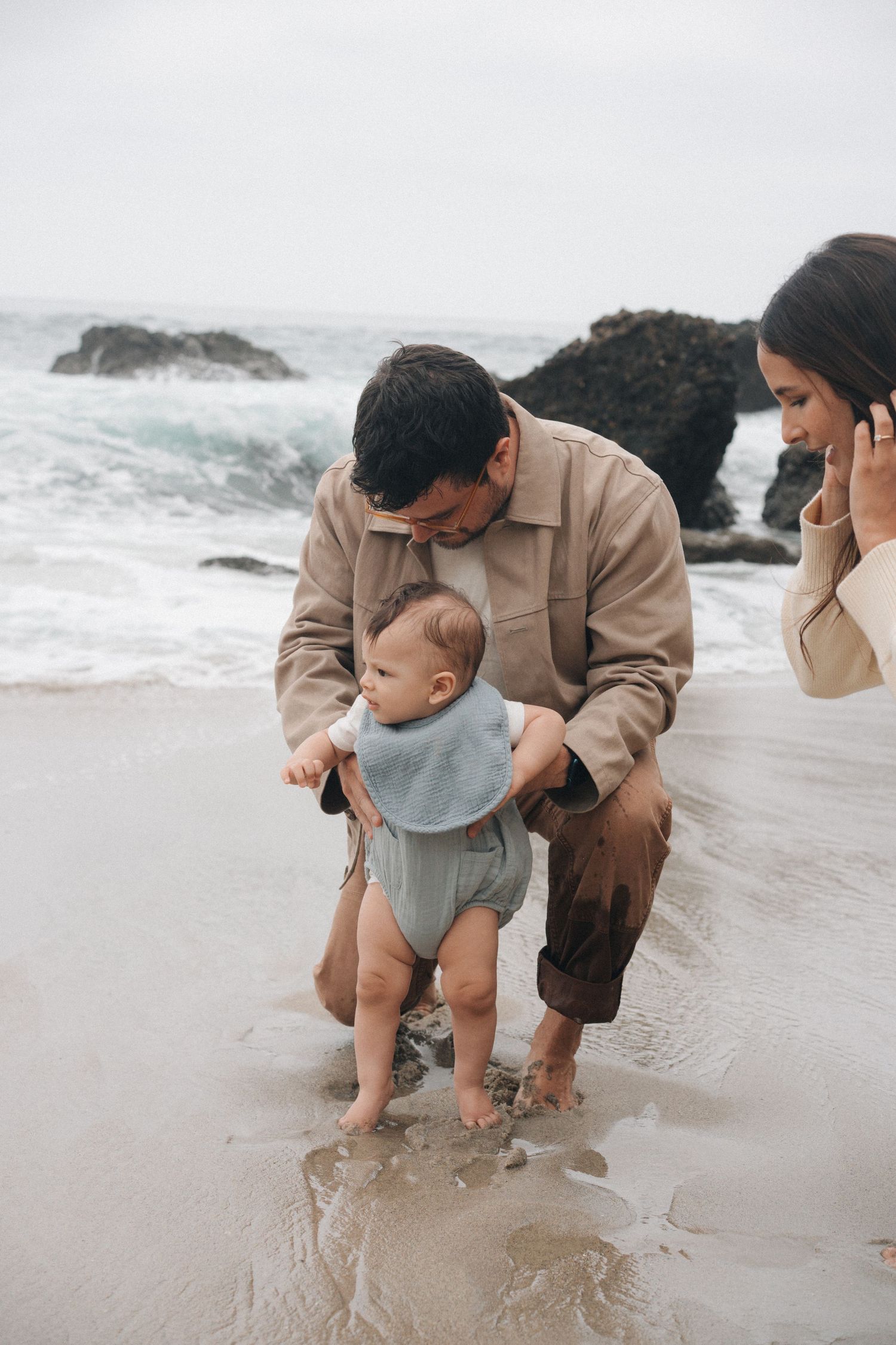 Parent helping toddler in gray onesie explore wet sand on a misty beach near ocean waves.