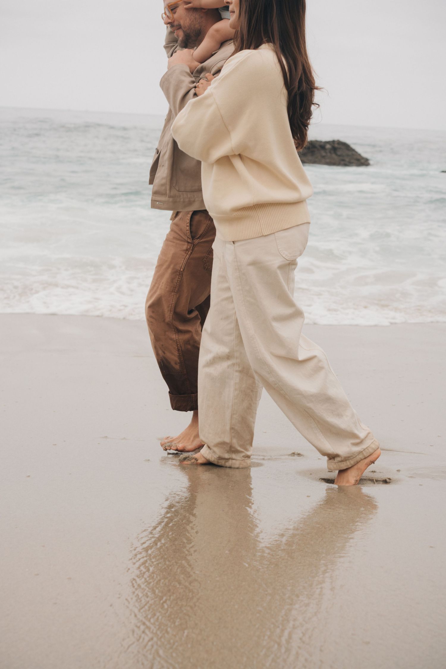 Two people stroll on a beach wearing neutral-colored casual clothing, their feet touching the wet sand and ocean waves.