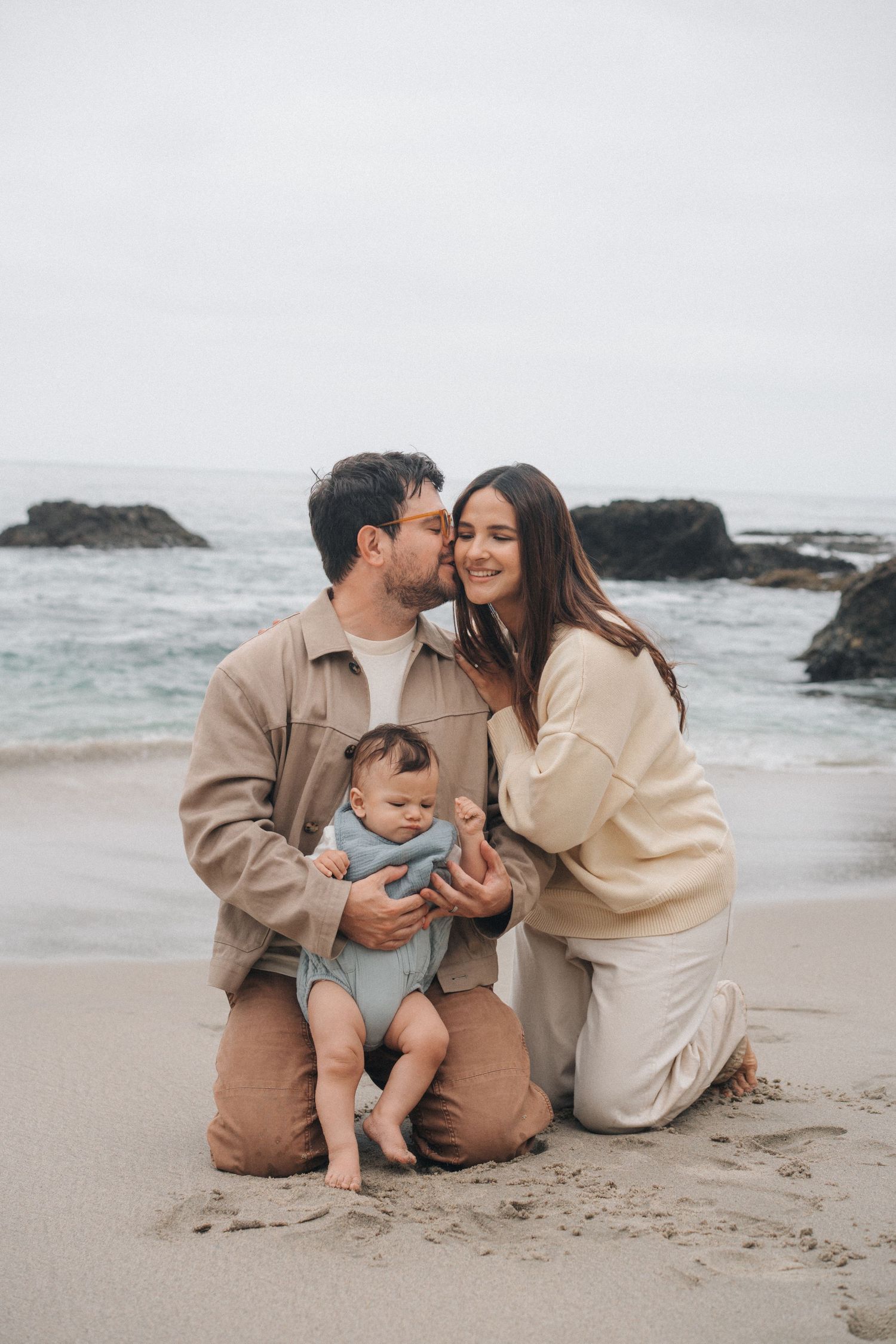 A family wearing matching beige outfits sits together on a cloudy beach with waves and rocks in the background.