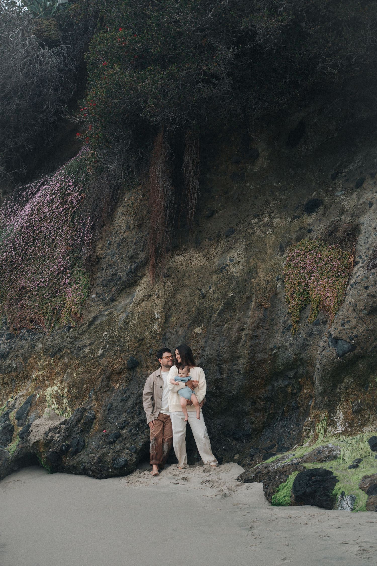 Couple poses together against dramatic rocky cliffs along a scenic beach at sunset.