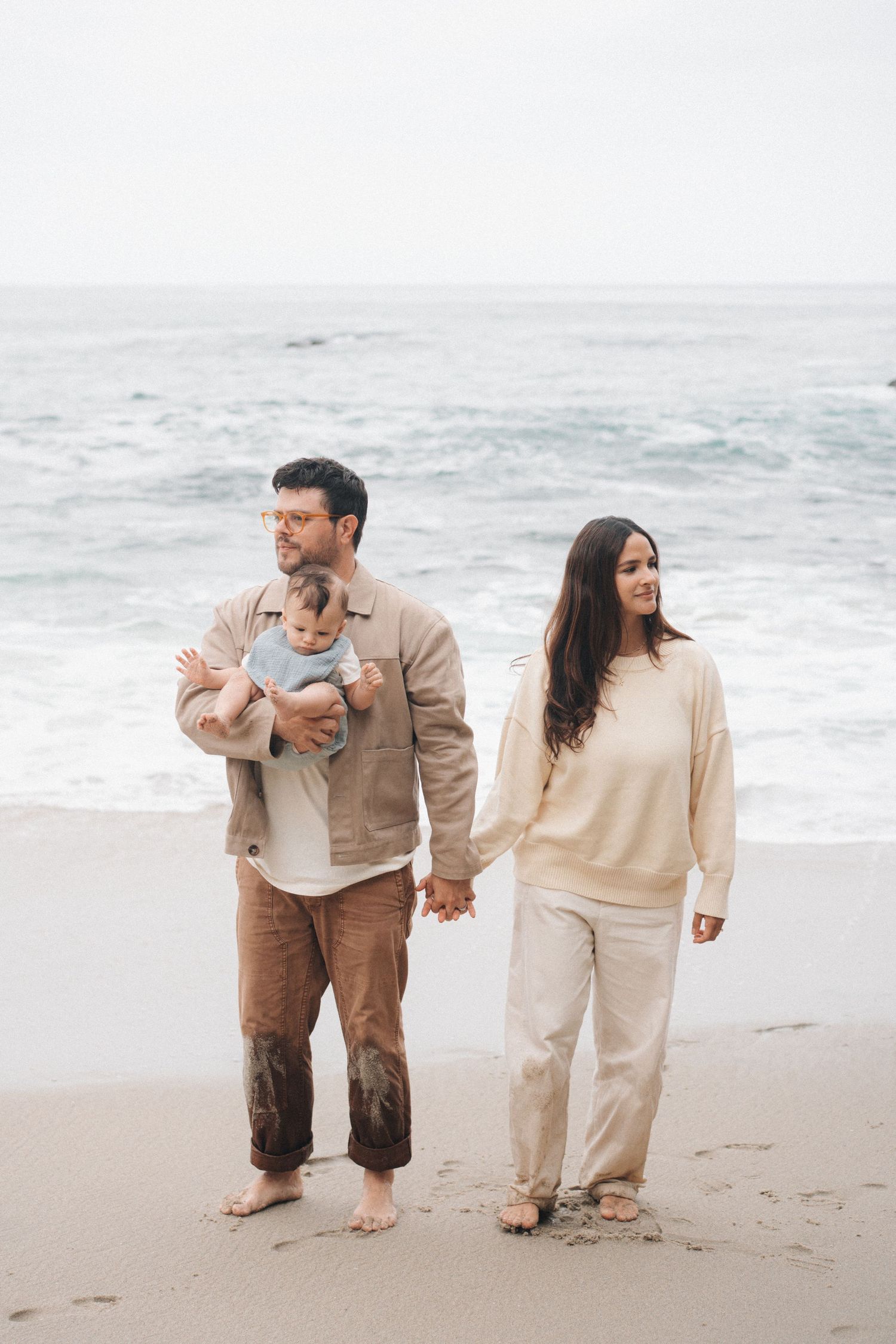 A couple in neutral-toned clothing stands together on a foggy beach while holding their baby.