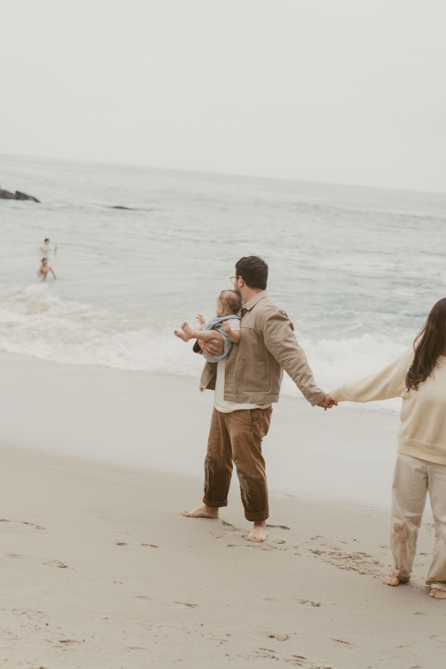 A small family walks hand in hand along a misty beach shore during an overcast day.