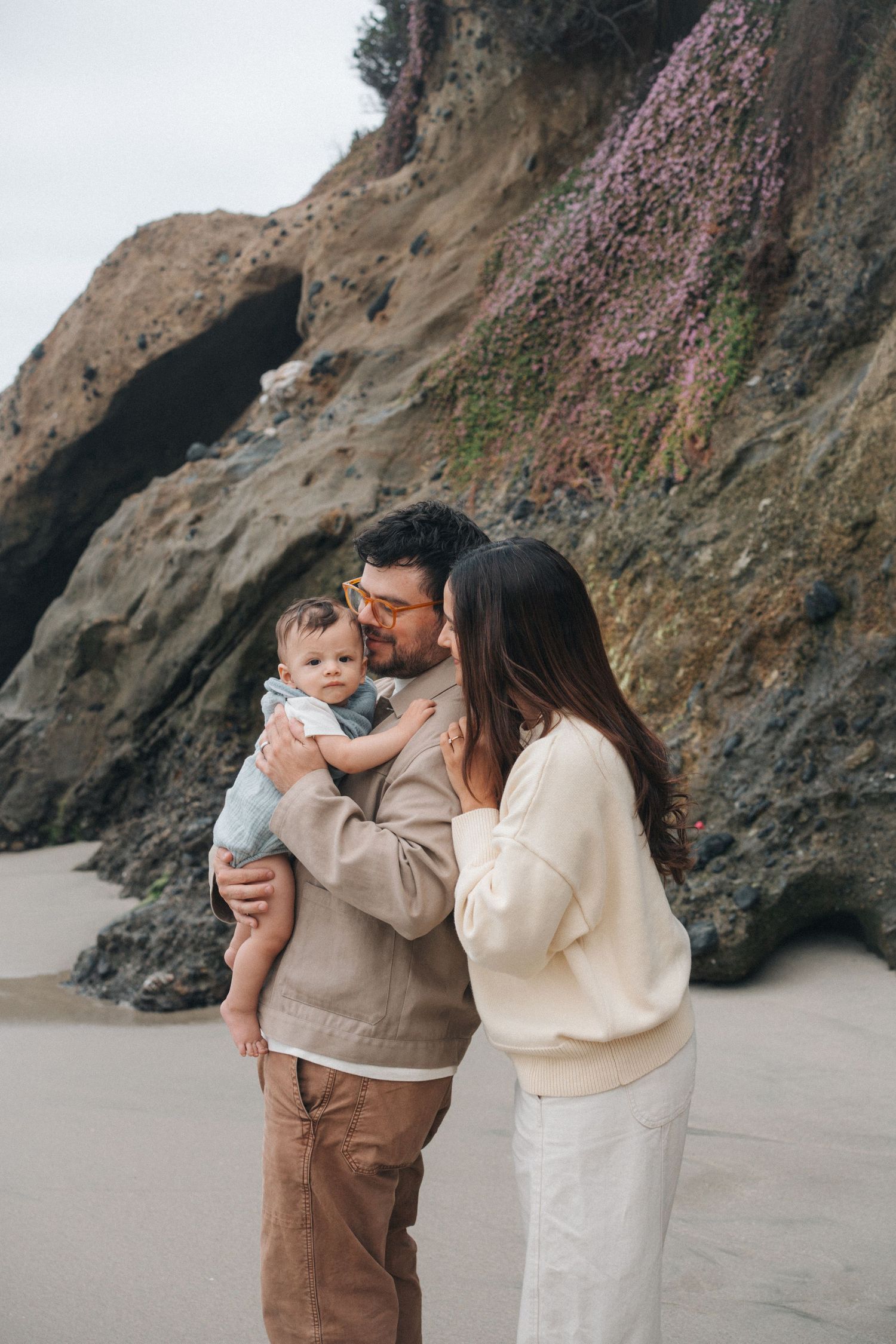 A family shares tender moments together on a scenic beach with rocky cliffs in the background.