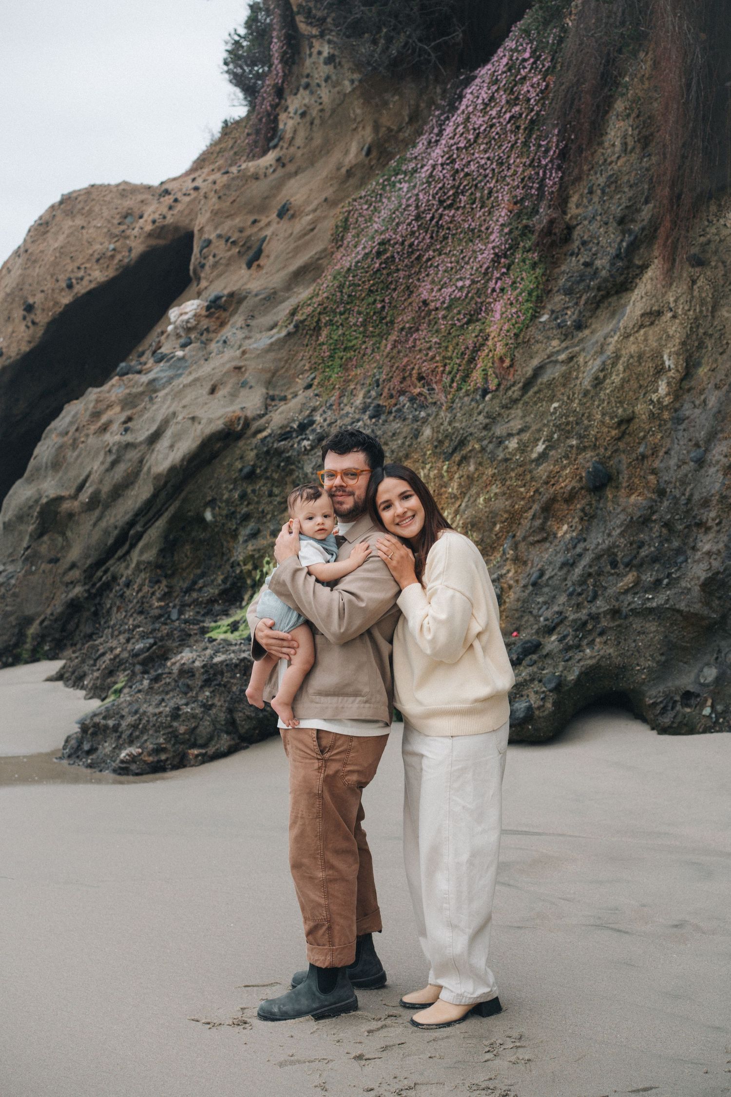 A family stands together on a sandy beach with dramatic rocky cliffs and pink wildflowers in the background.