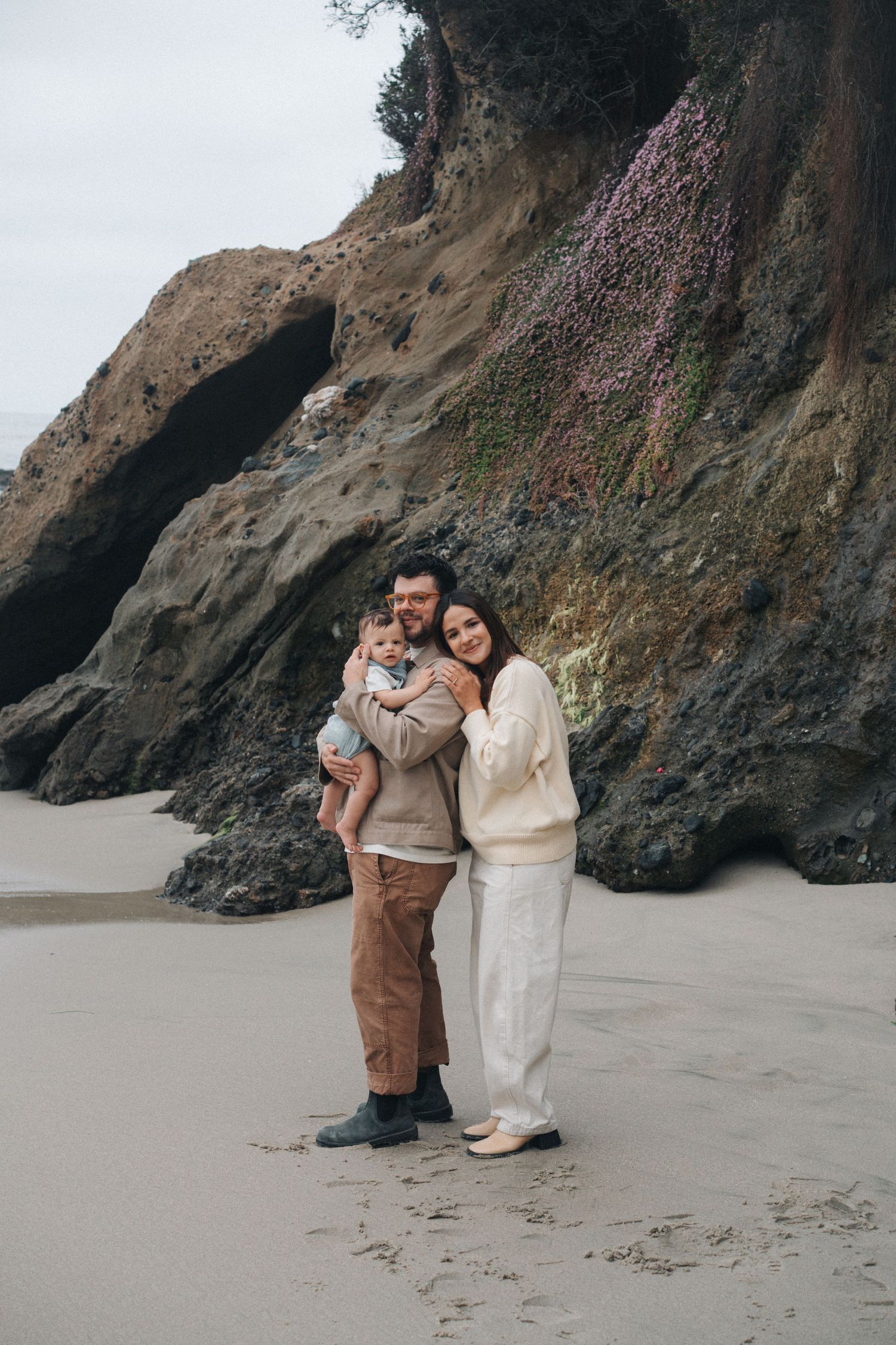 A family in cozy sweaters poses together on a moody beach with dramatic cliffs and rock formations in the background.