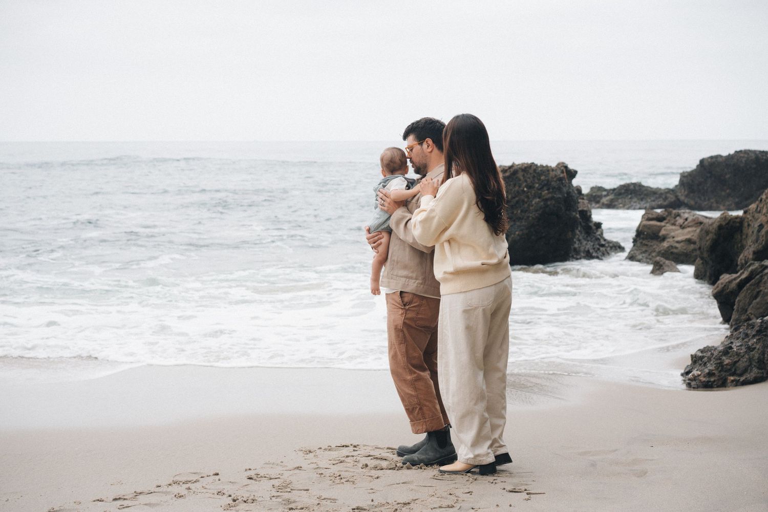 A couple embraces on a scenic beach with rocky cliffs and crashing waves in the background.