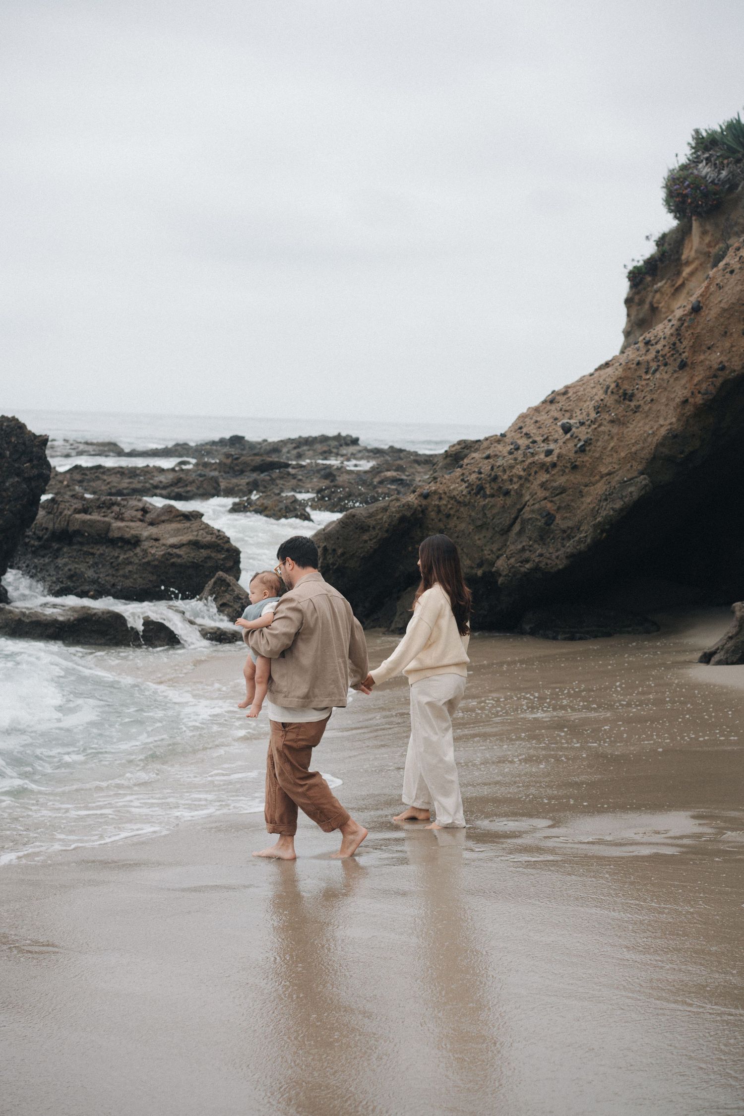 Couple walking along misty beach with rocky cliffs and crashing waves in the background.