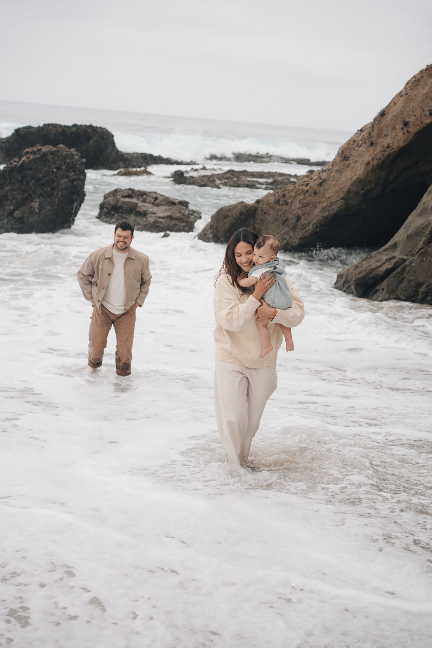 A couple in light beige outfits plays together at a rocky beach as ocean waves crash against the shore.