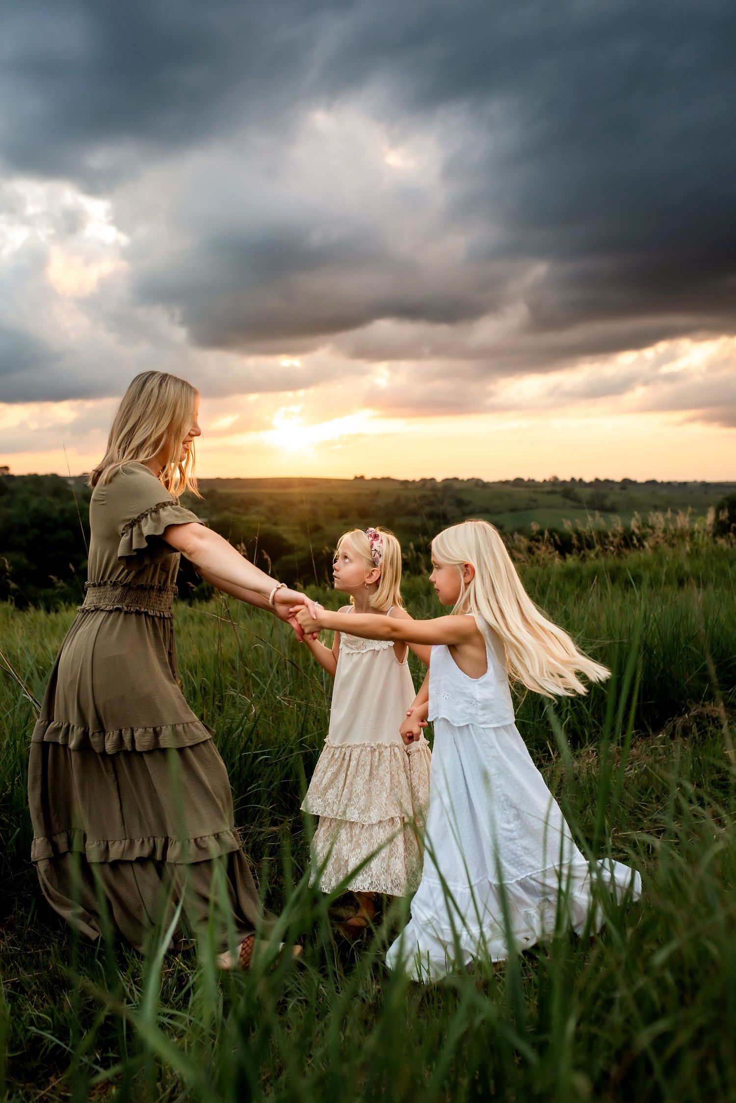 Three figures dance together in a grassy field at sunset beneath dramatic storm clouds.