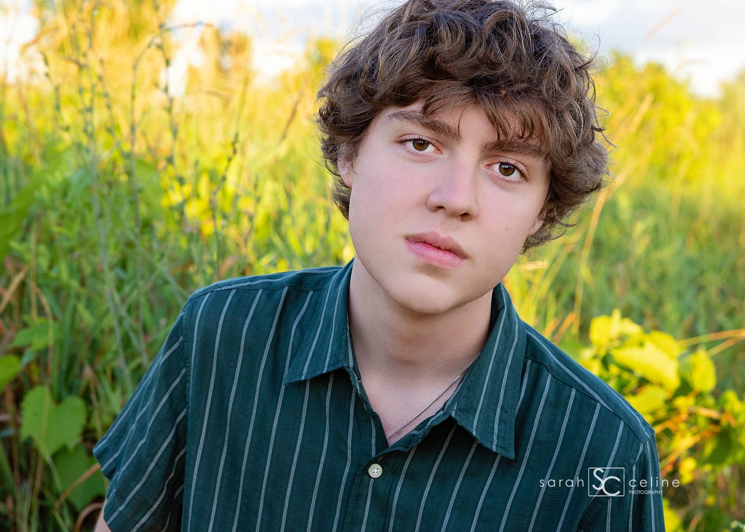A person in a dark green striped button-down shirt poses outdoors in a sunlit field with yellow wildflowers.