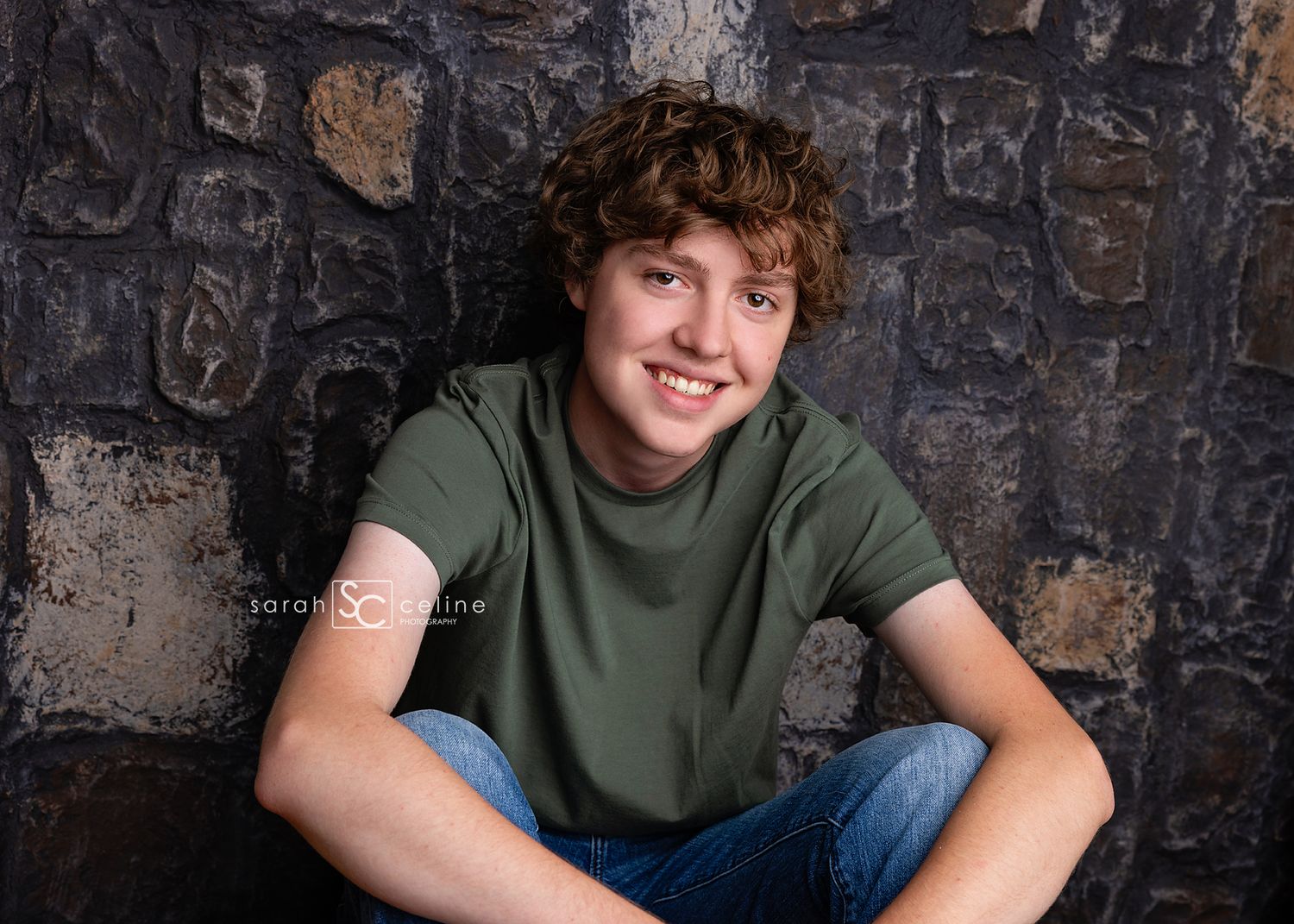 A teen in a green t-shirt and jeans sits casually against a textured stone wall background.