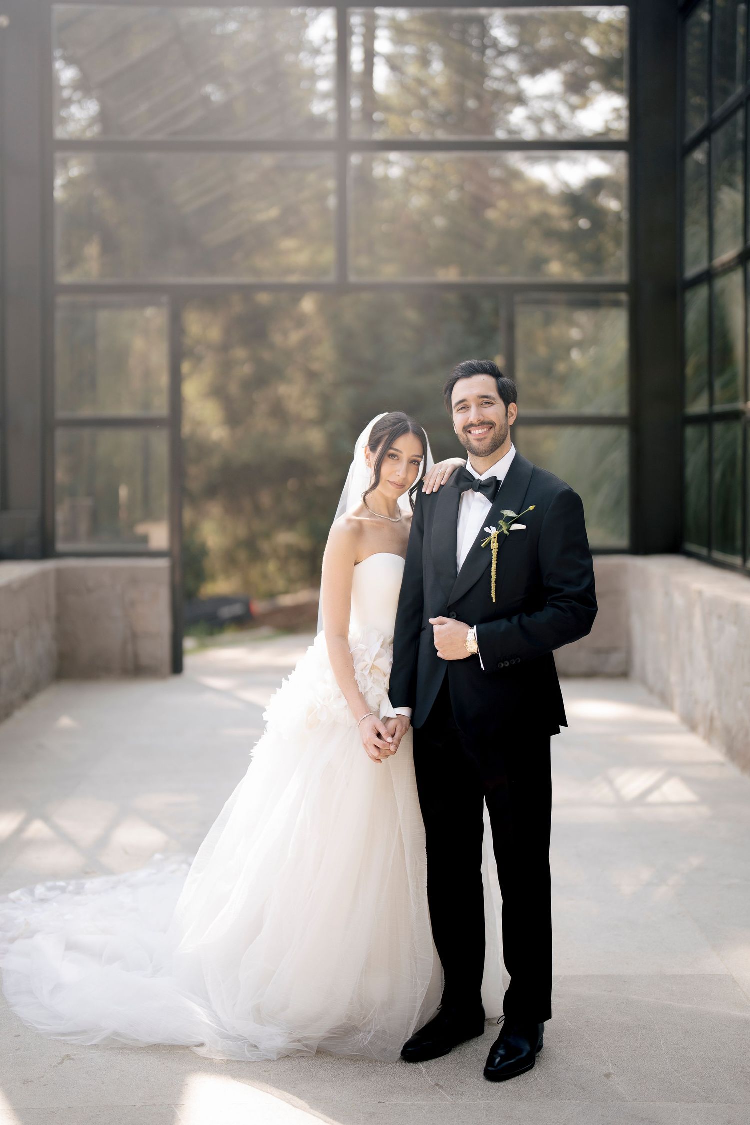 Elegant bride in white lace gown and groom in classic tuxedo pose together in sunlit glass corridor at wedding venue.