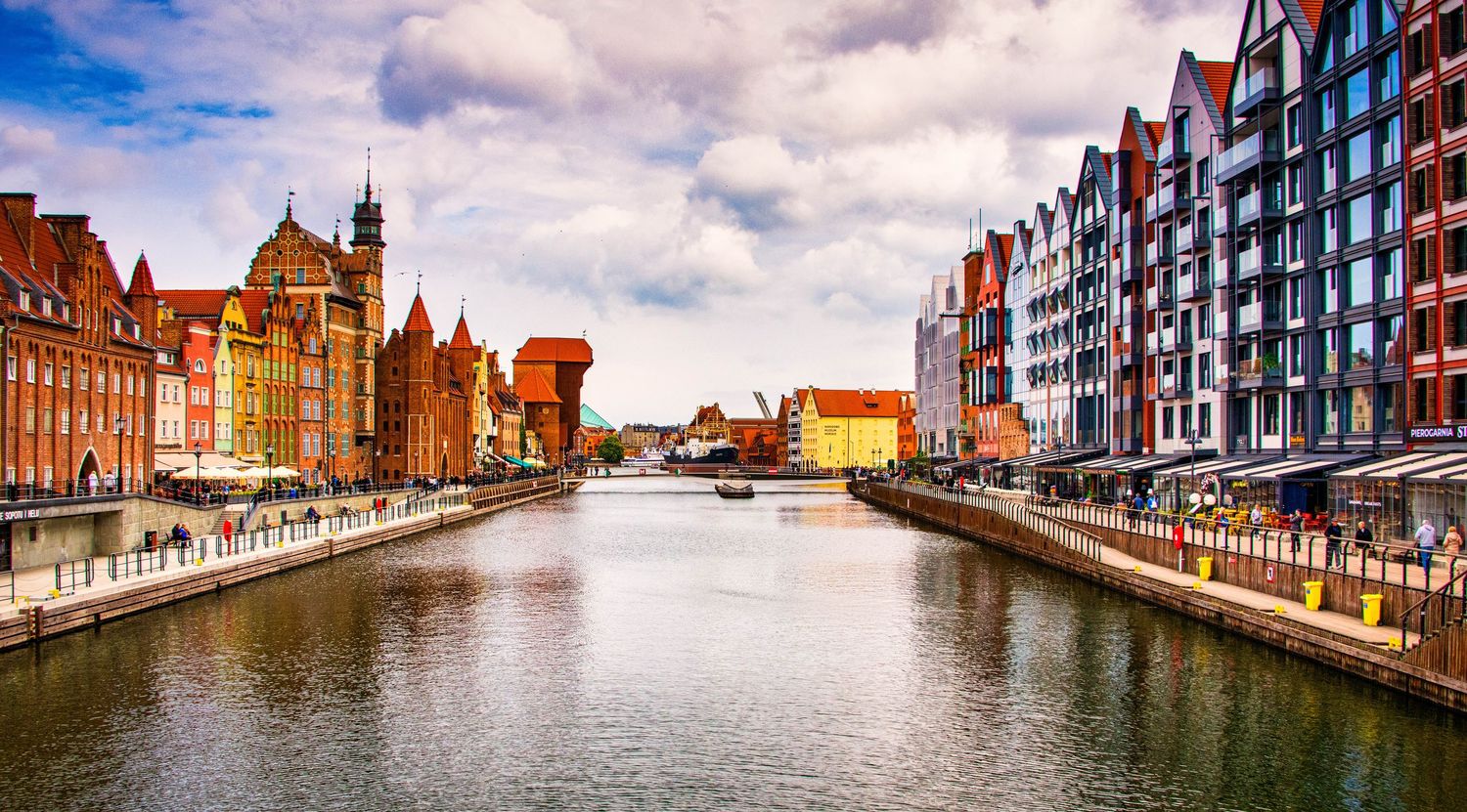 Panoramic cityscape of Gdansk's colorful waterfront buildings reflecting in the harbor under blue skies.