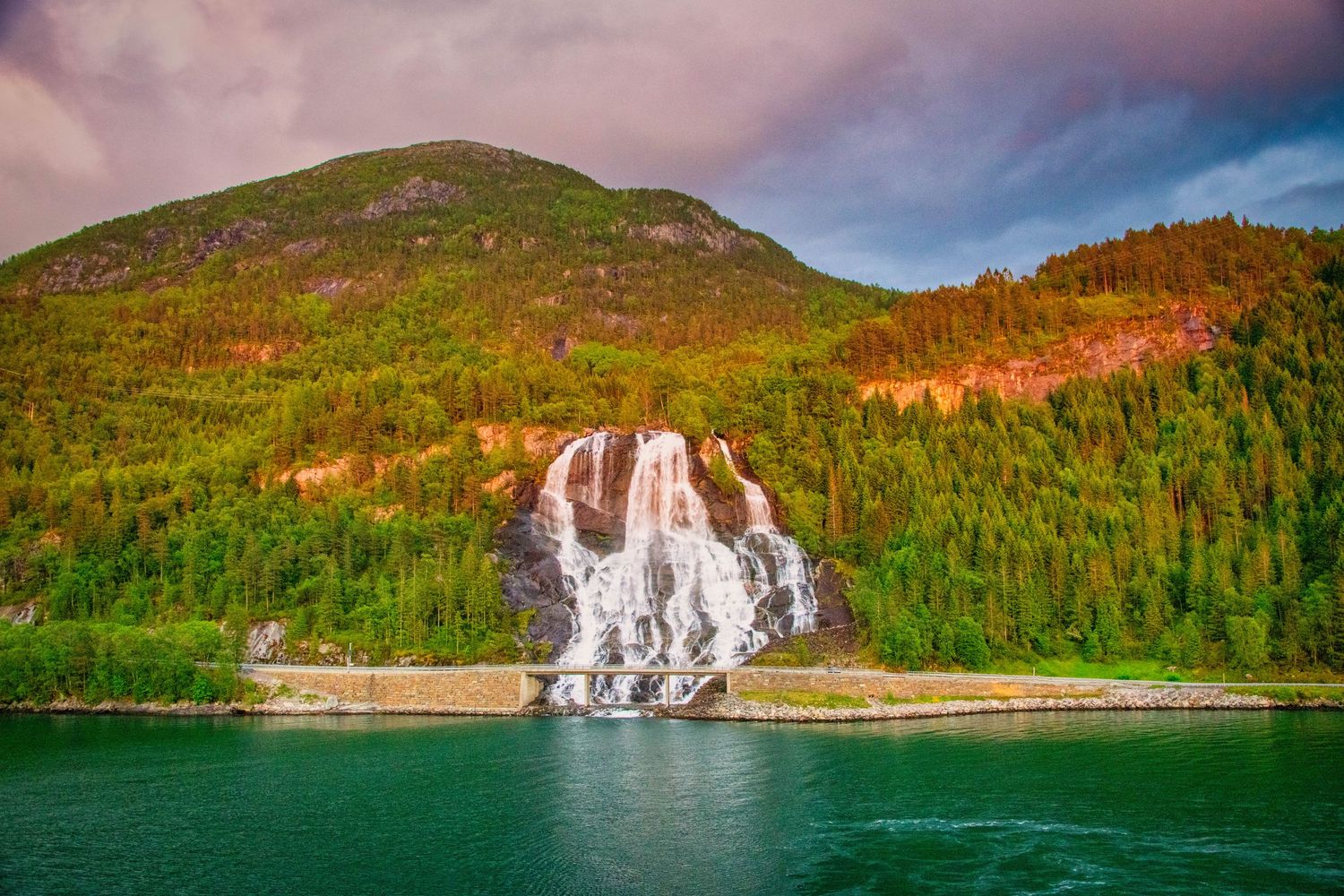 Cascading waterfalls flow down green mountainsides into a pristine lake surrounded by forest in a scenic landscape.