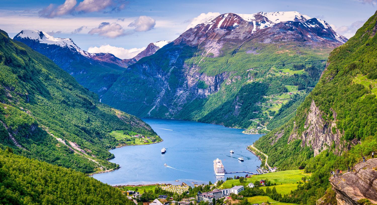 Panoramic view of Norwegian fjords with snow-capped mountains and lush green valleys.