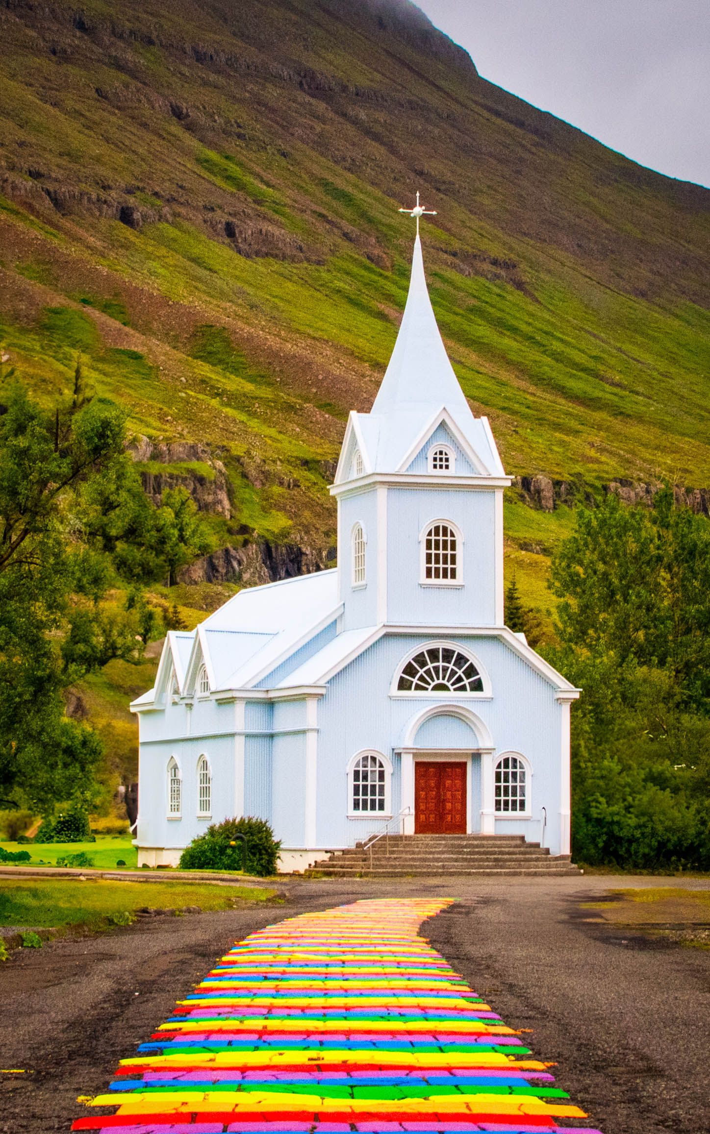 White wooden church with colorful rainbow painted steps leading to entrance against mountain backdrop.