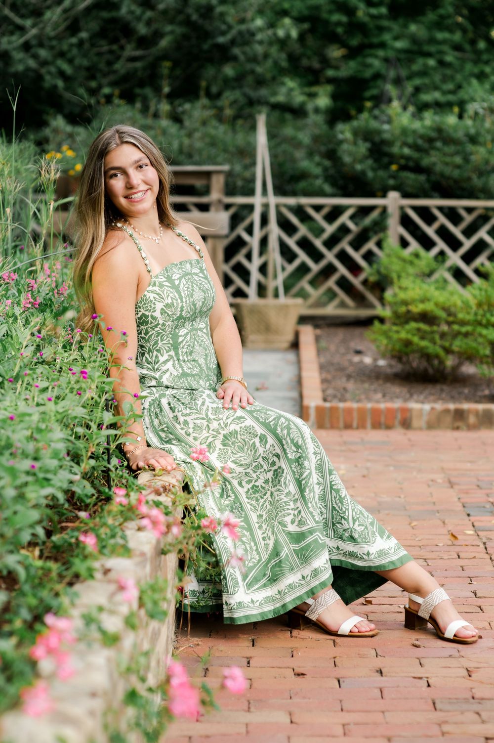 A person wearing a green and white patterned sundress sits on a brick path in a garden setting with flowers and lattice.