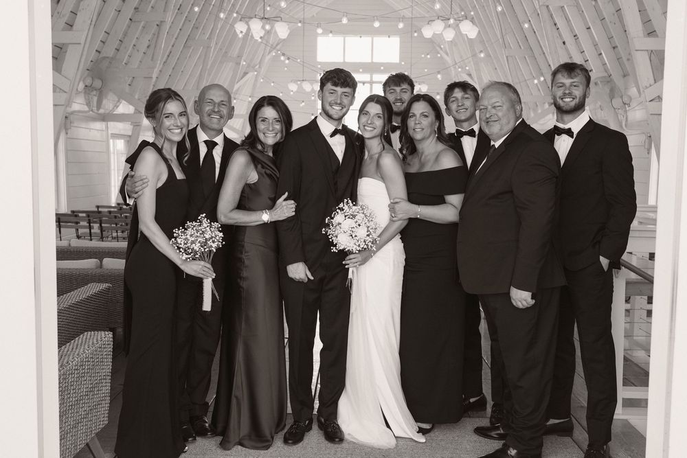 Wedding party members in formal attire pose together in a rustic venue with string lights overhead.