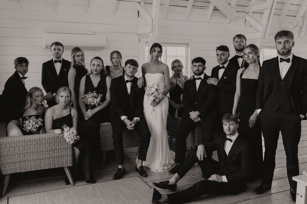 Wedding party poses together in black formal attire against a white backdrop.