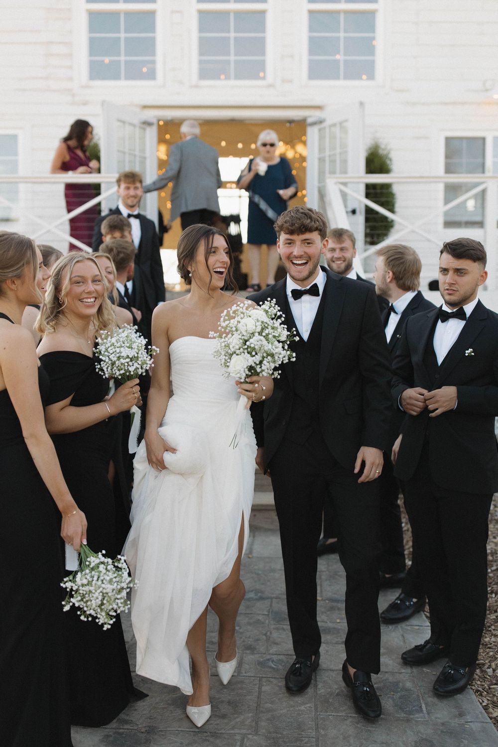 Wedding party members laugh and interact on a paved outdoor area near a white building.