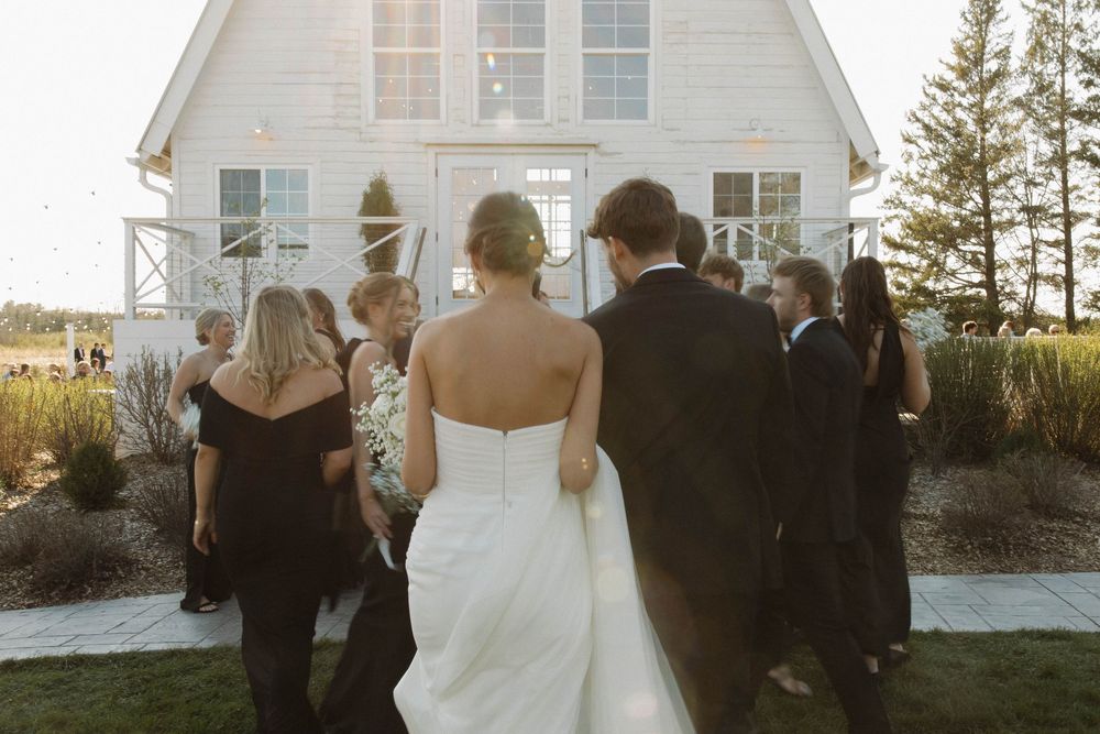 Wedding party gathers outside a white chapel-style building for formal photos.