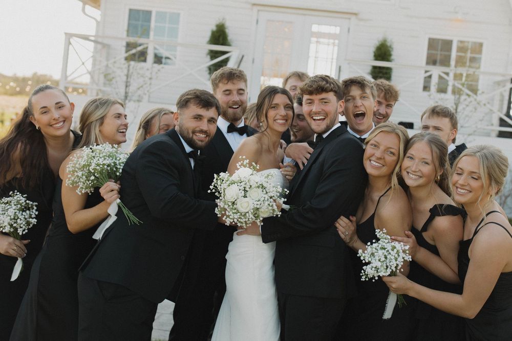 Bridal party members in black dresses and suits pose together outdoors near a white building.