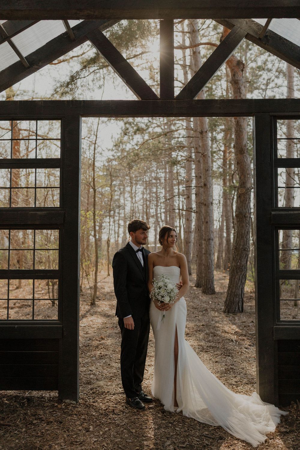 A couple shares an intimate moment inside a wooden chapel structure surrounded by tall pine trees.