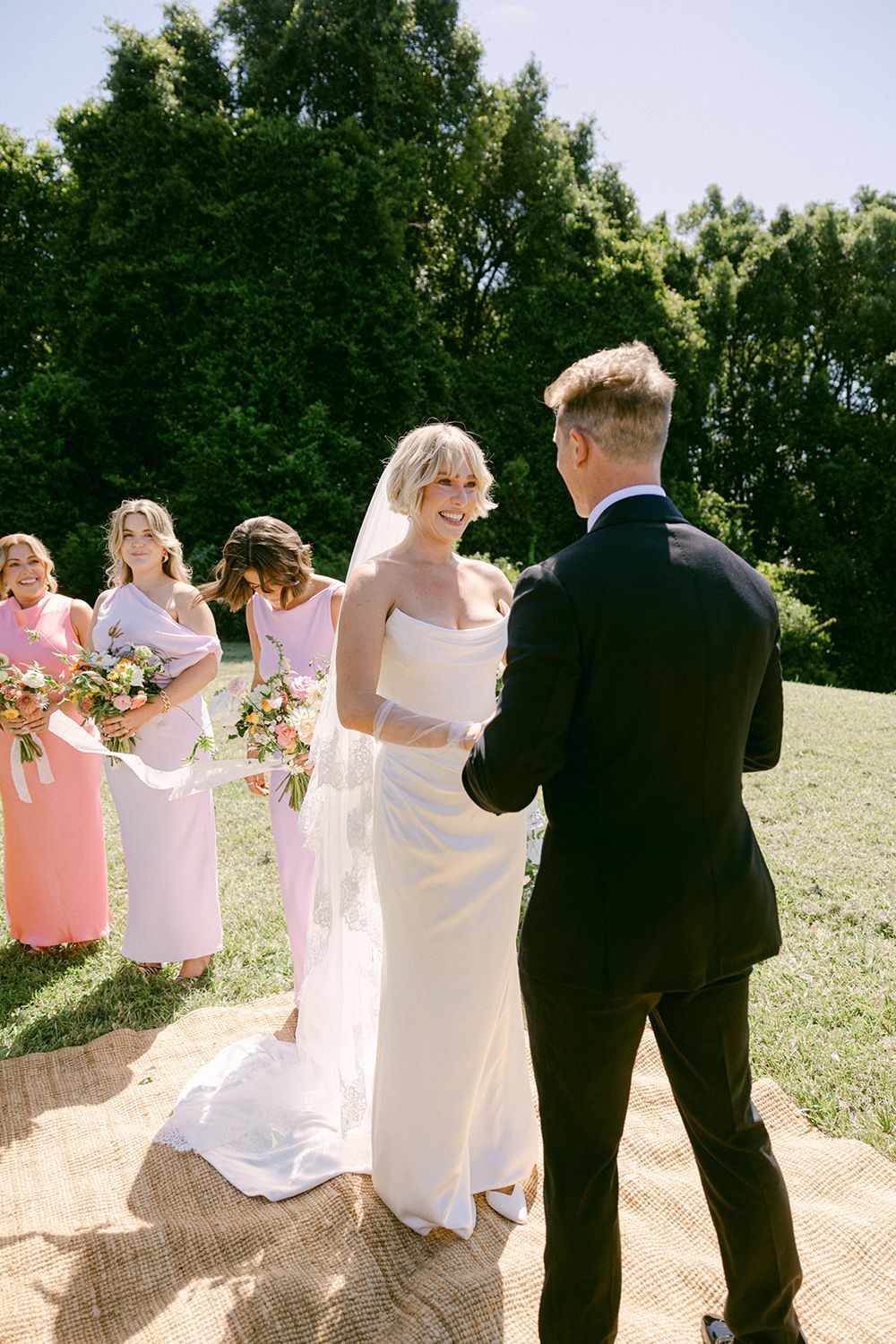A wedding ceremony takes place outdoors with bridesmaids in pink dresses holding bouquets on a sunny summer day.