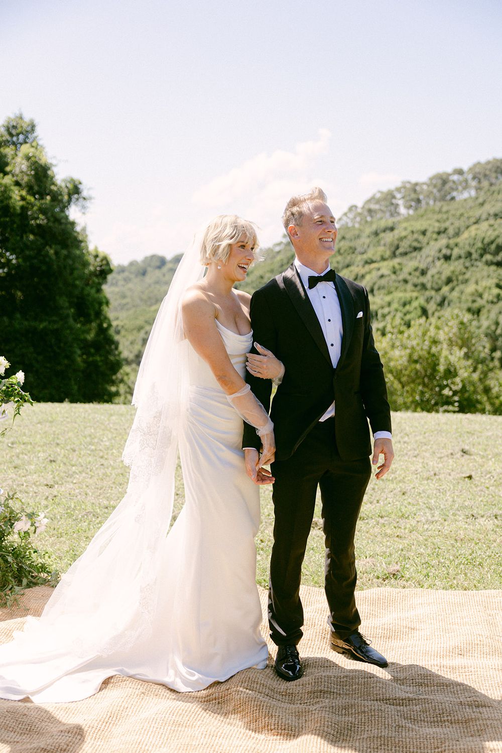 Newlywed couple in formal wedding attire stand together outdoors on a sunny day with green hills in the background.