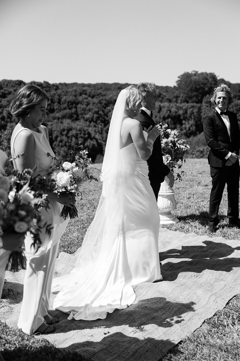 Black and white photo of an outdoor wedding ceremony with a bride in a flowing white dress and attendants holding bouquets.