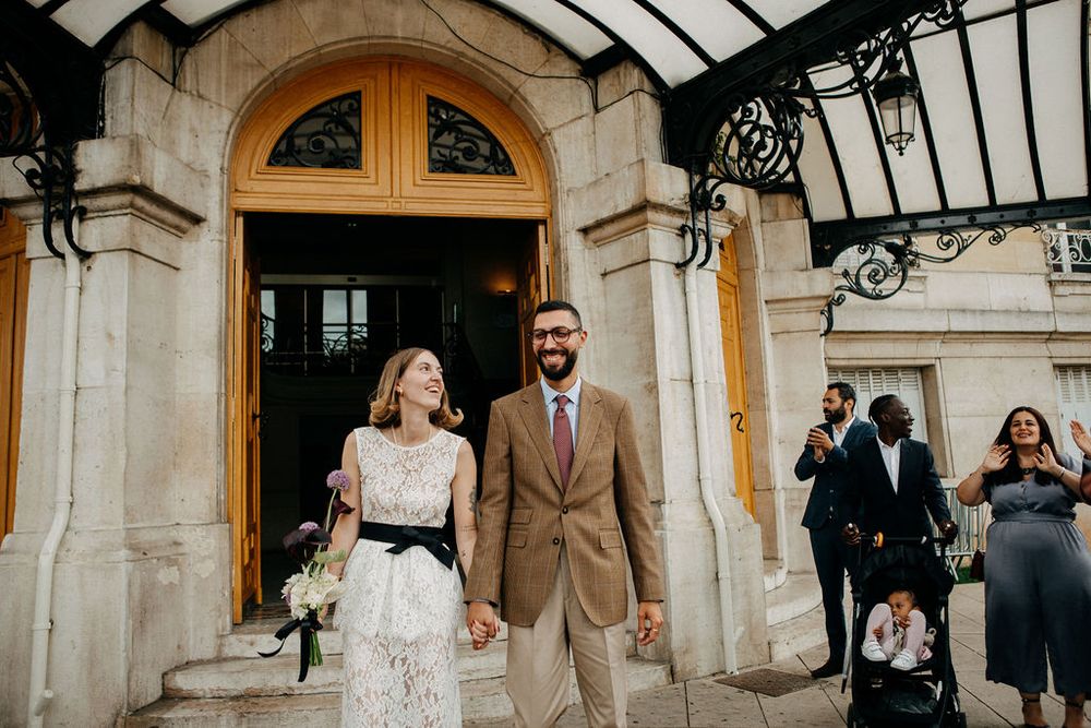 Newlywed couple holding hands and smiling as they exit a grand building with ornate doors after their wedding ceremony.