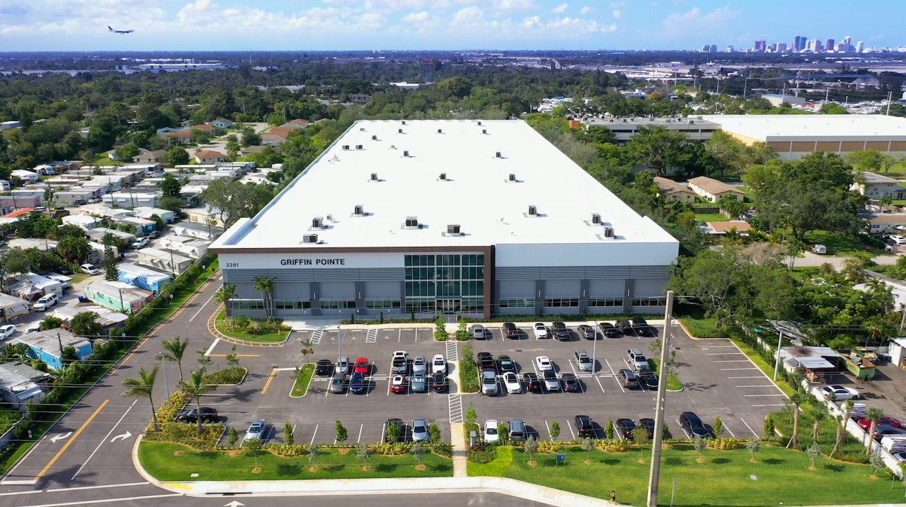 Aerial view of modern industrial warehouse facility with large parking lot, palm-lined entrance, and urban skyline in the background.