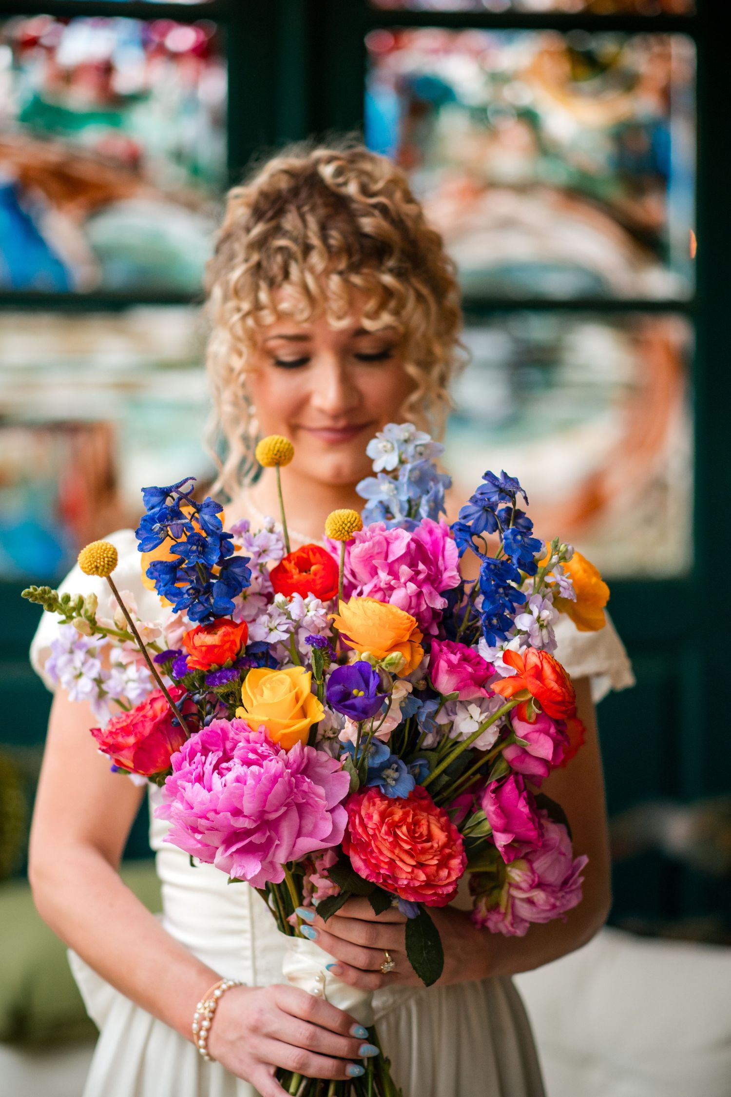 A colorful spring wedding bouquet with vibrant peonies, orange roses, and blue delphinium flowers held against a blurred background.