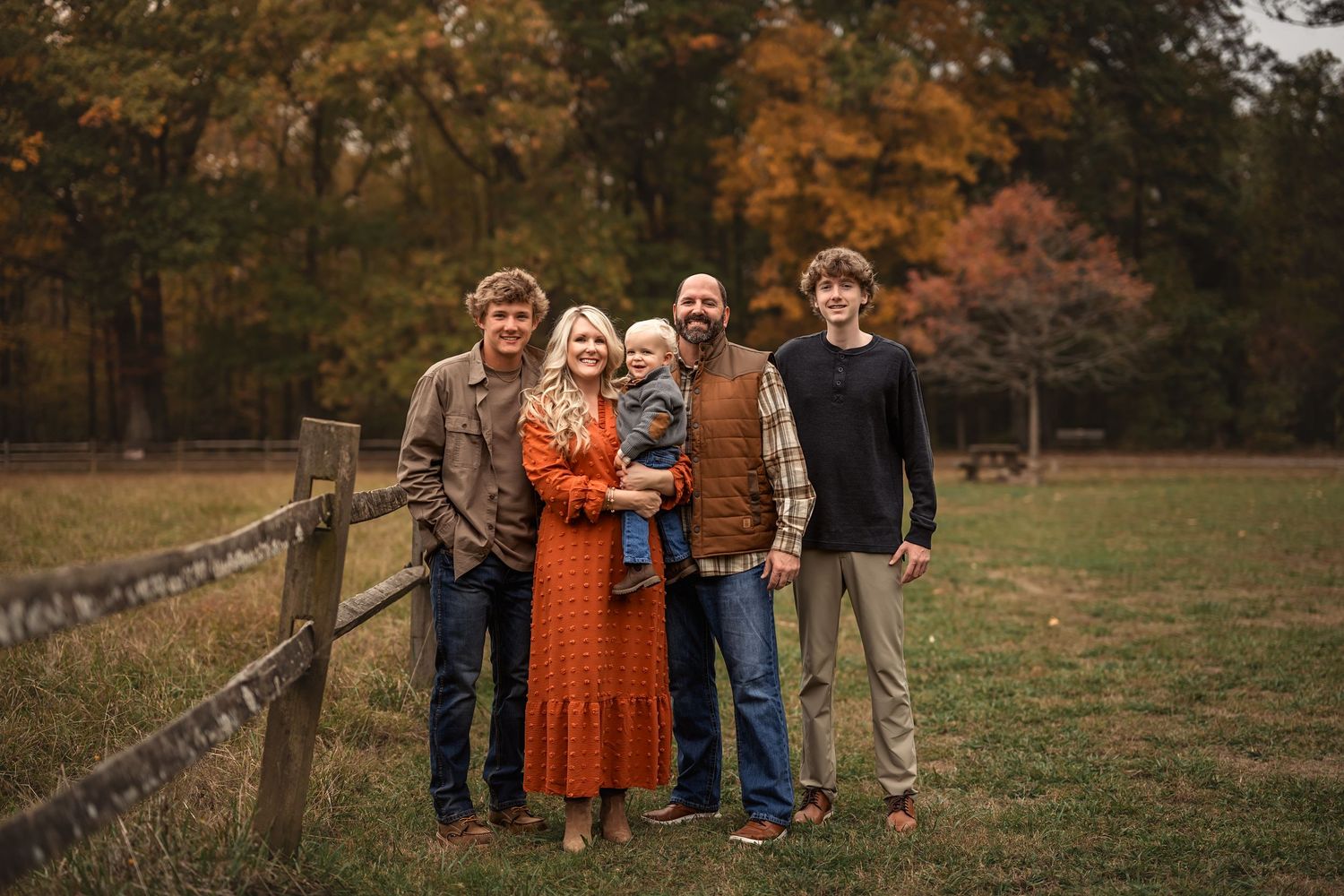 A family stands together by a rustic wooden fence during autumn with fall foliage in the background.